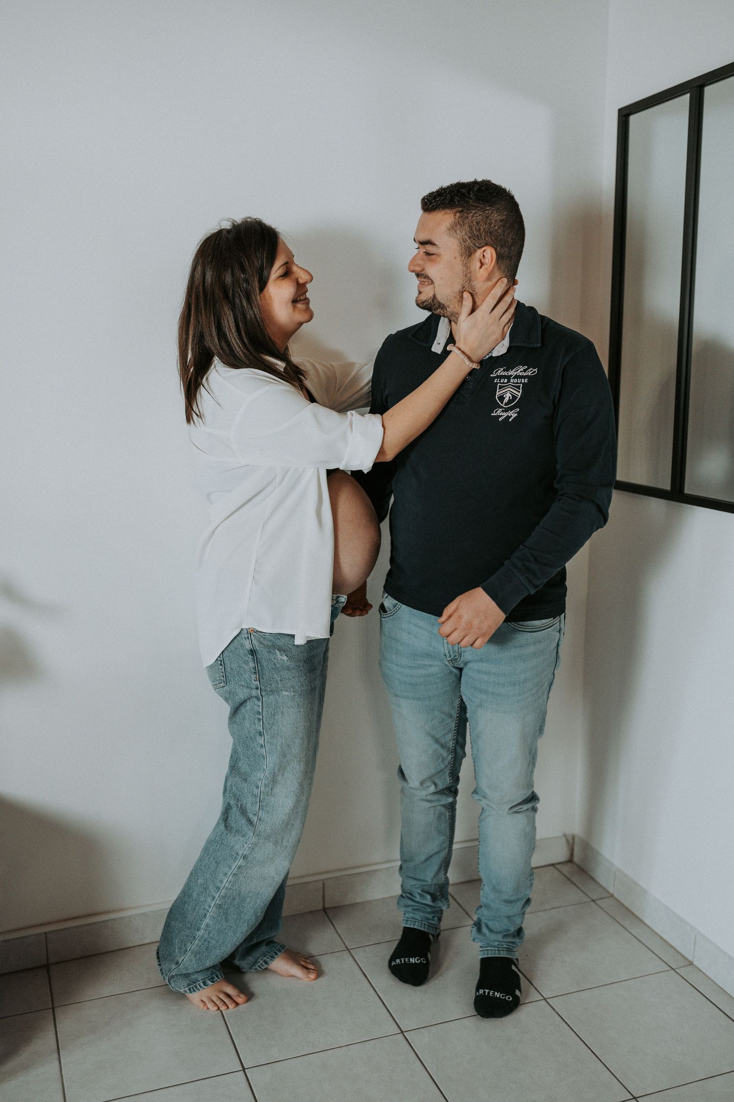 A pregnant woman in white shirt and jeans tenderly holds a man's face while they share a loving moment indoors.