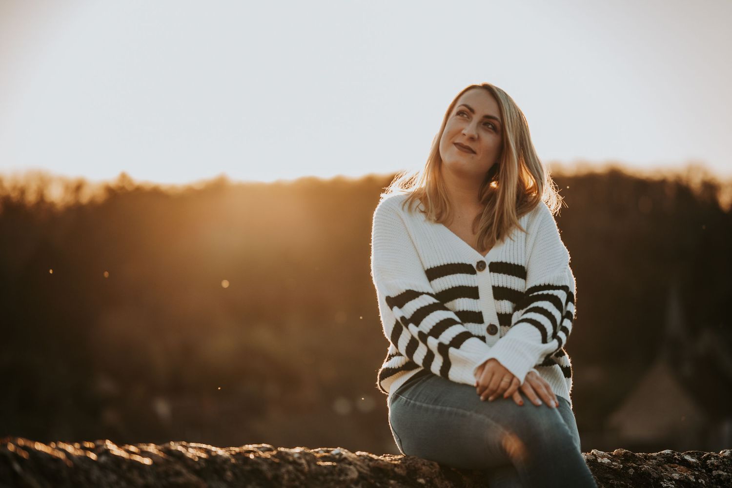 Woman in striped sweater sitting outdoors at golden hour, bathed in warm sunset light with a dreamy, contemplative expression.
