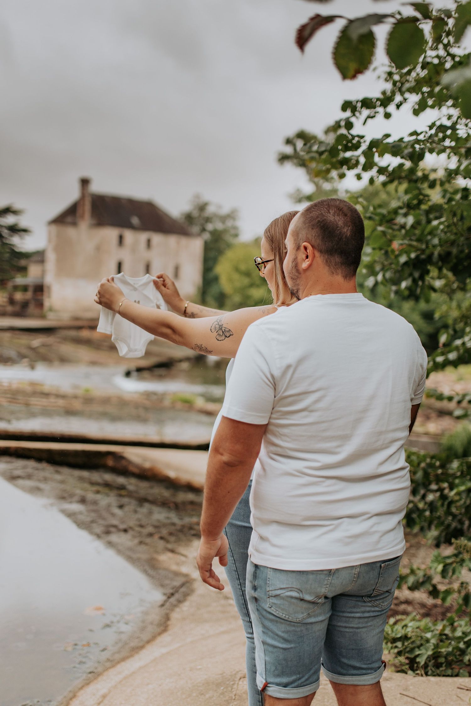 A couple taking scenic photos by a waterfront with buildings in the background on a cloudy day.