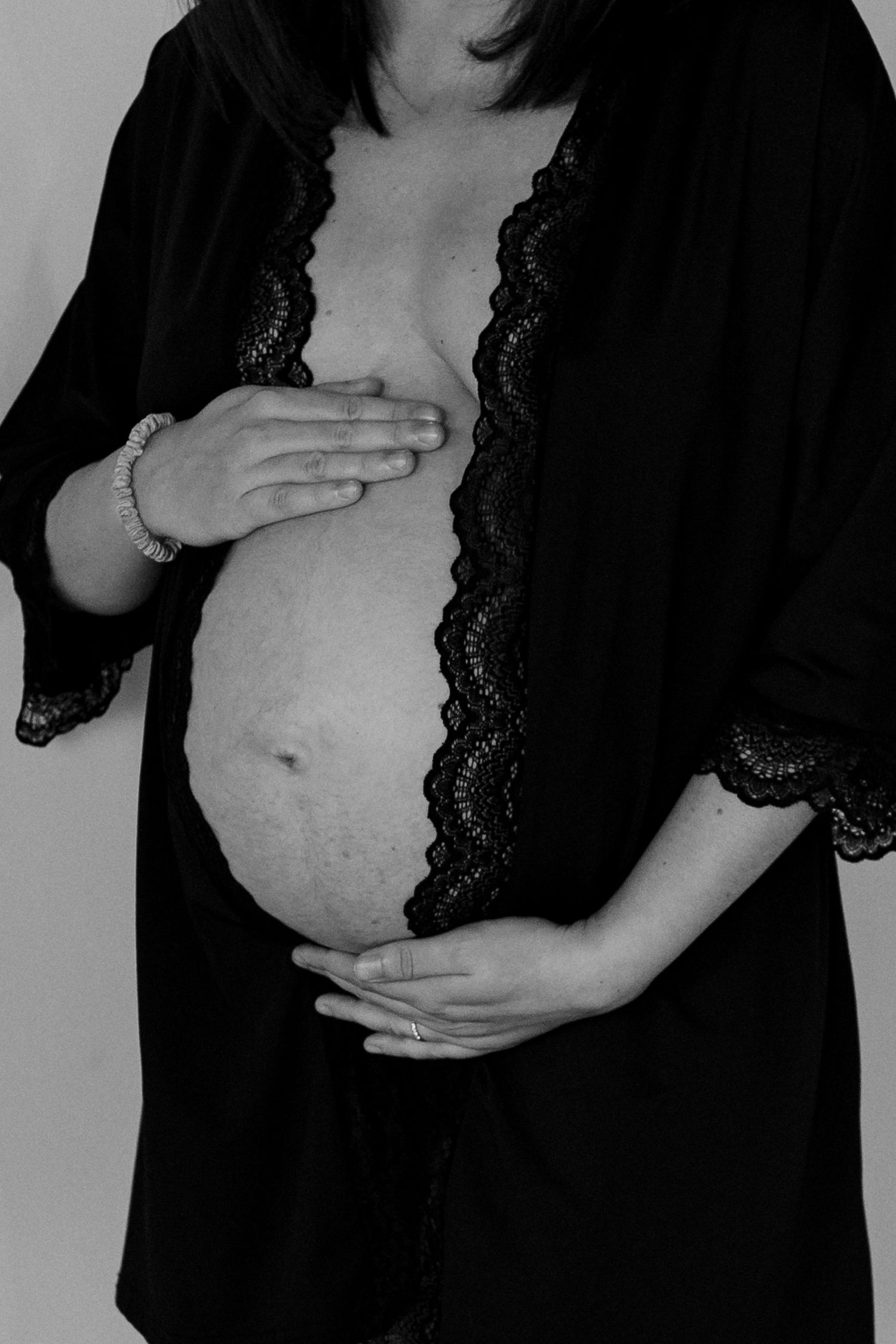 Black and white maternity photo showing pregnant belly with hands cradling bump, wearing lace-trimmed robe.