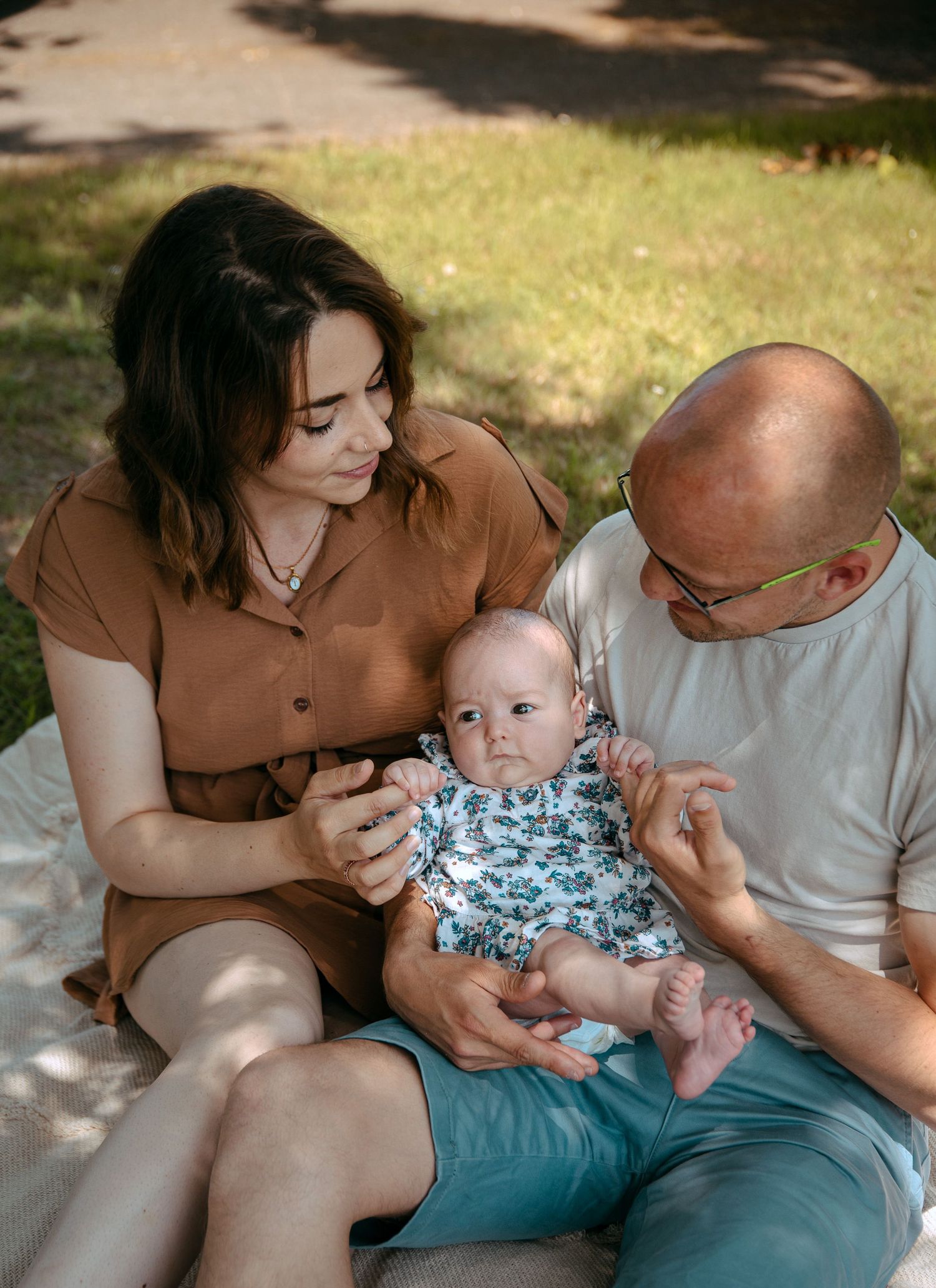 A small baby wearing a floral outfit sits outdoors with adults on a sunny summer day.