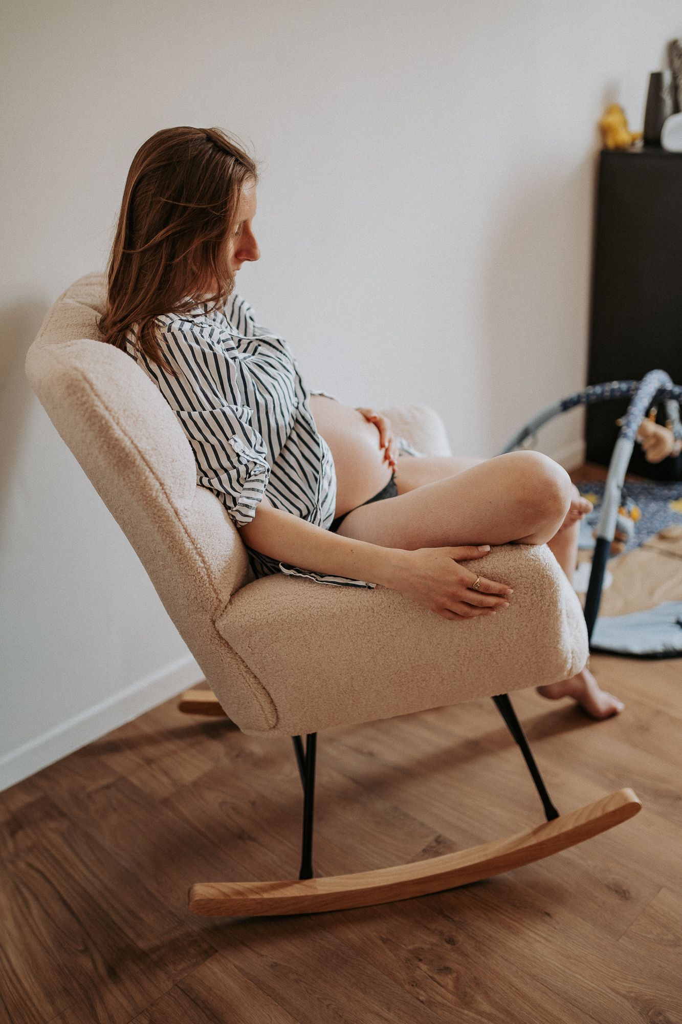 Pregnant woman sitting in a beige rocking chair, cradling her baby bump while looking to the side in a nursery room.