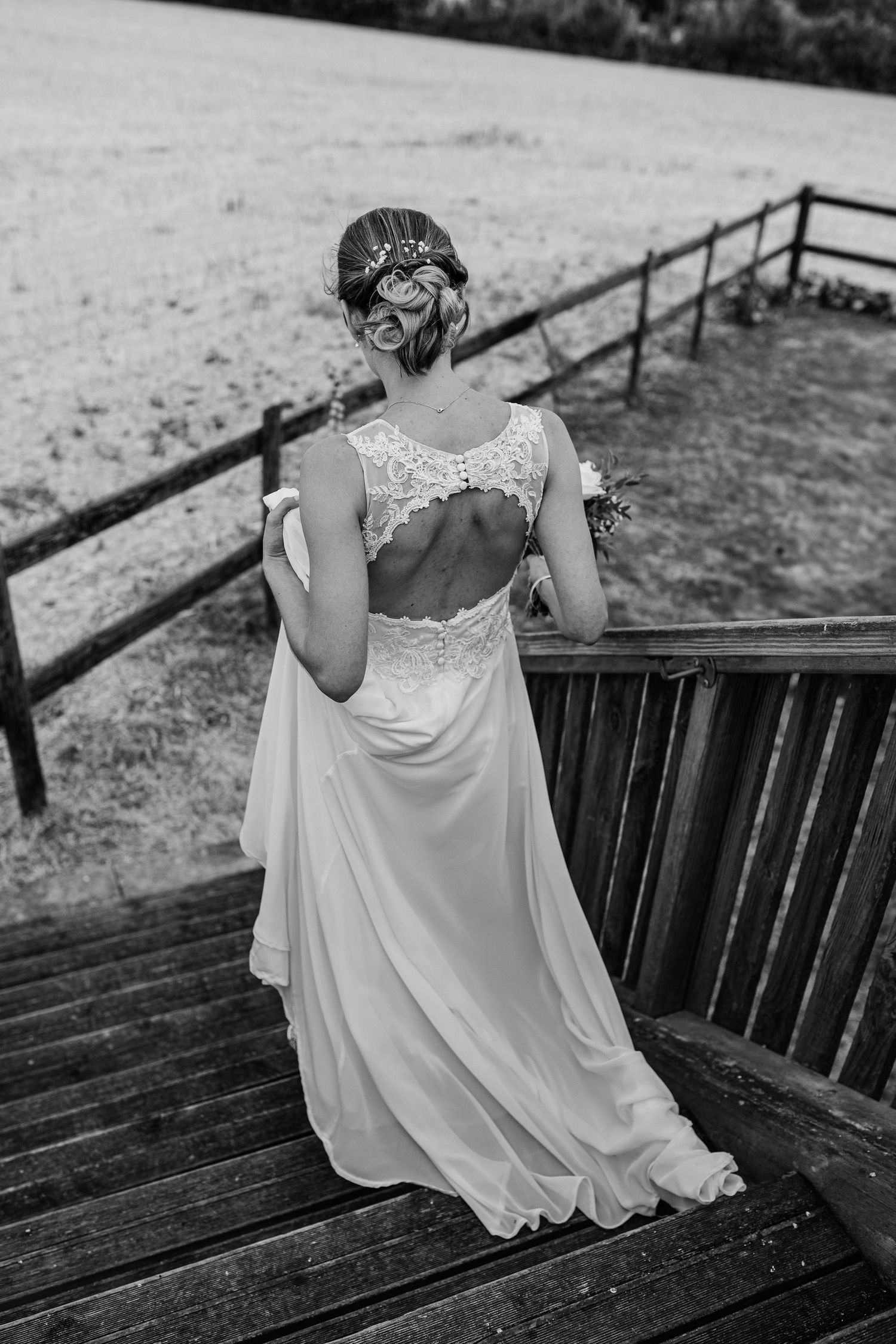 Back view of a flowing white wedding dress with lace details as someone sits on wooden steps outdoors.
