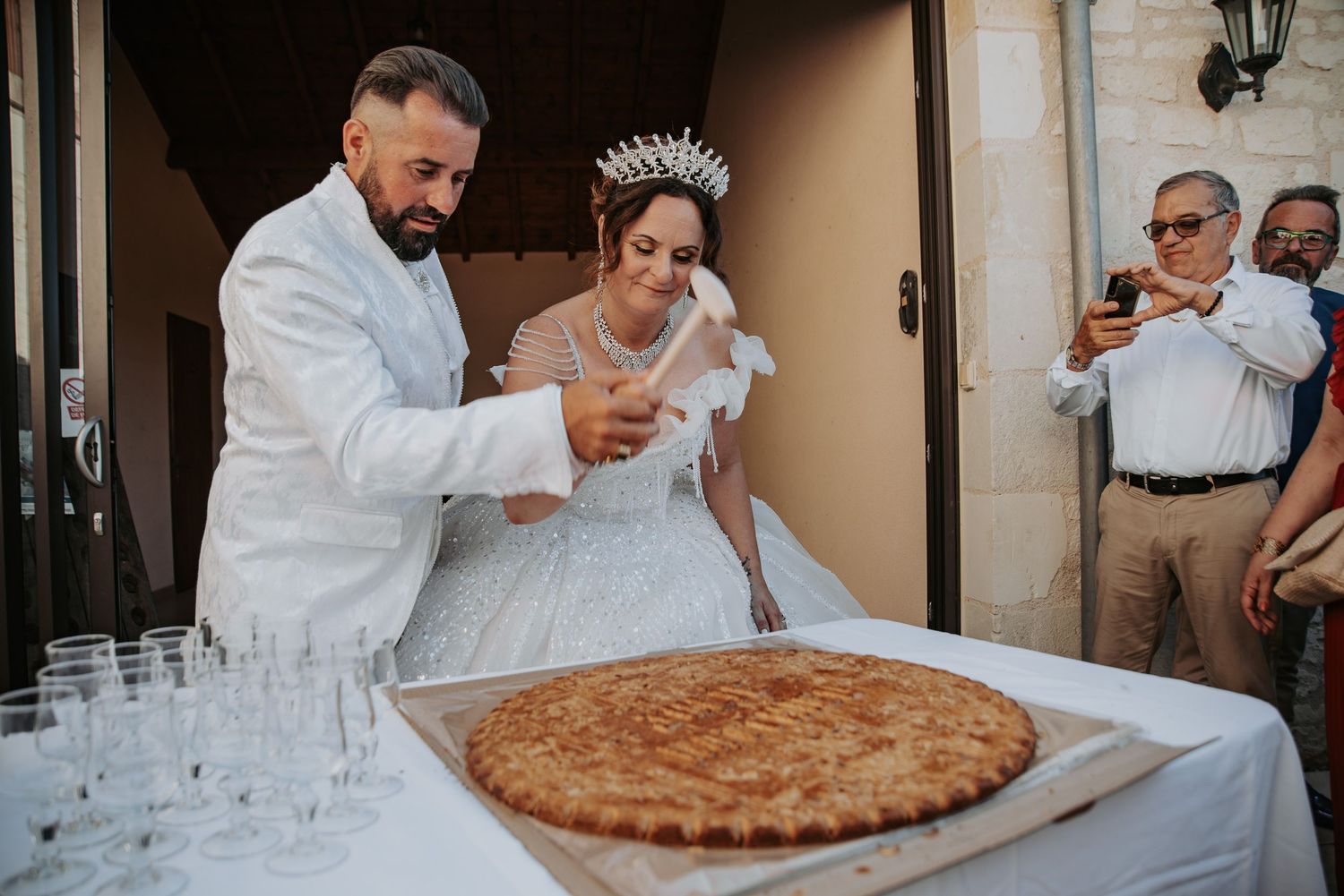 A wedding couple cuts into a large round pizza at their reception while guests watch and champagne glasses await.