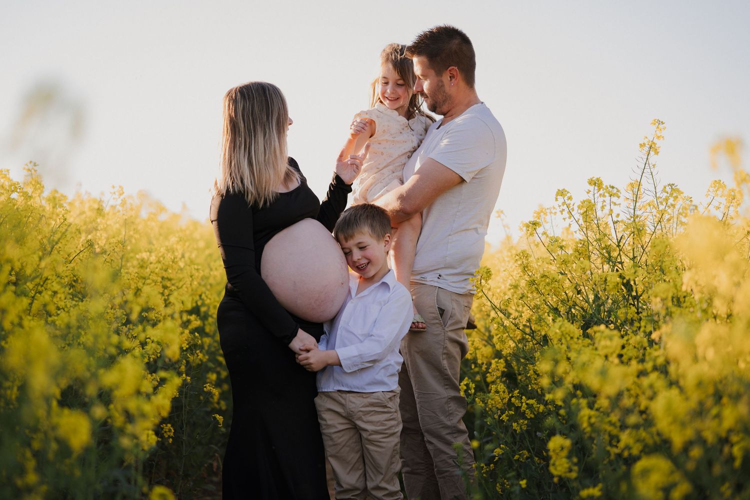 Pregnant woman with family in yellow canola field at golden hour, children touching baby bump.