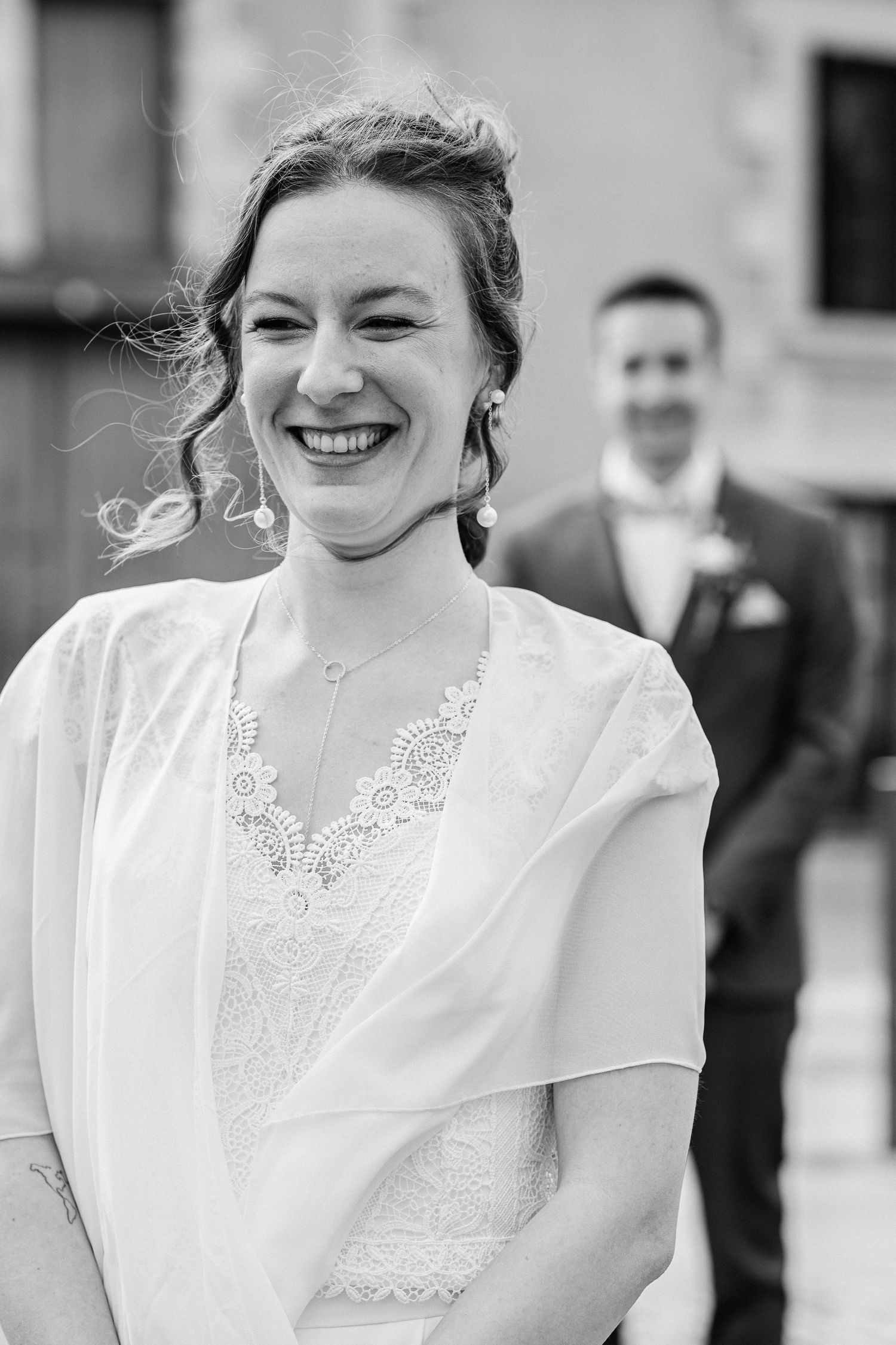 Black and white photo of a smiling bride in lace dress with groom blurred in background at outdoor wedding.