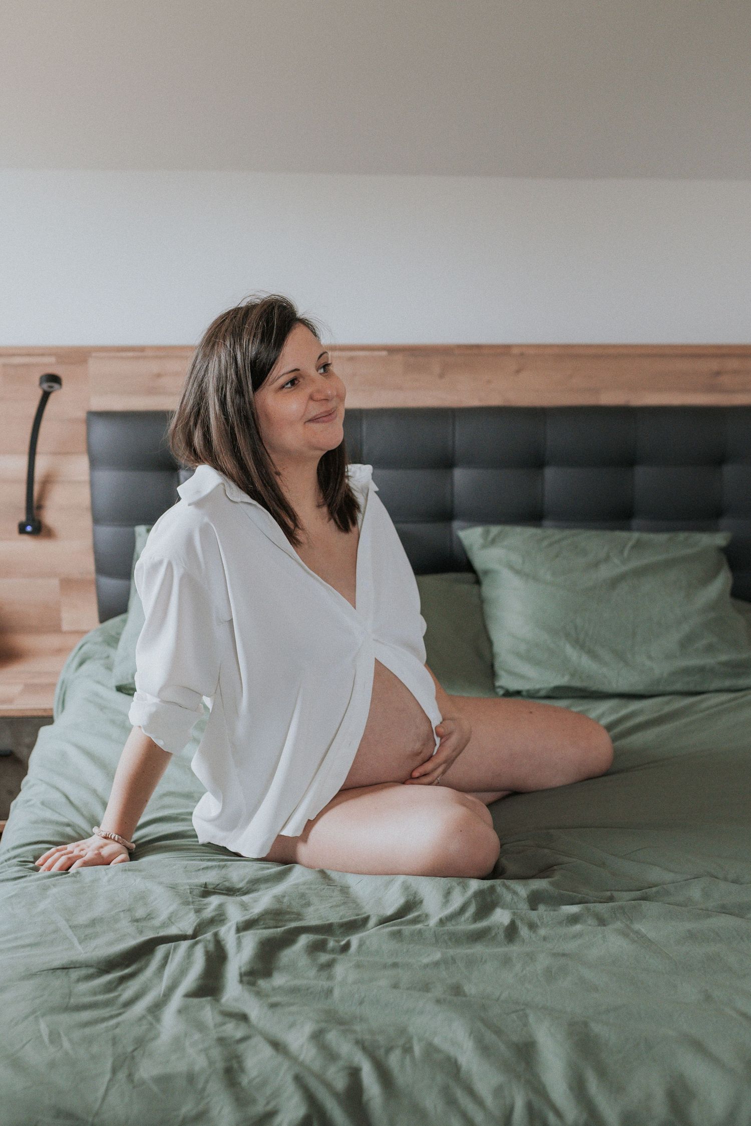 A person in a white button-down shirt relaxes on a bed with sage green bedding in a bright, airy bedroom setting.