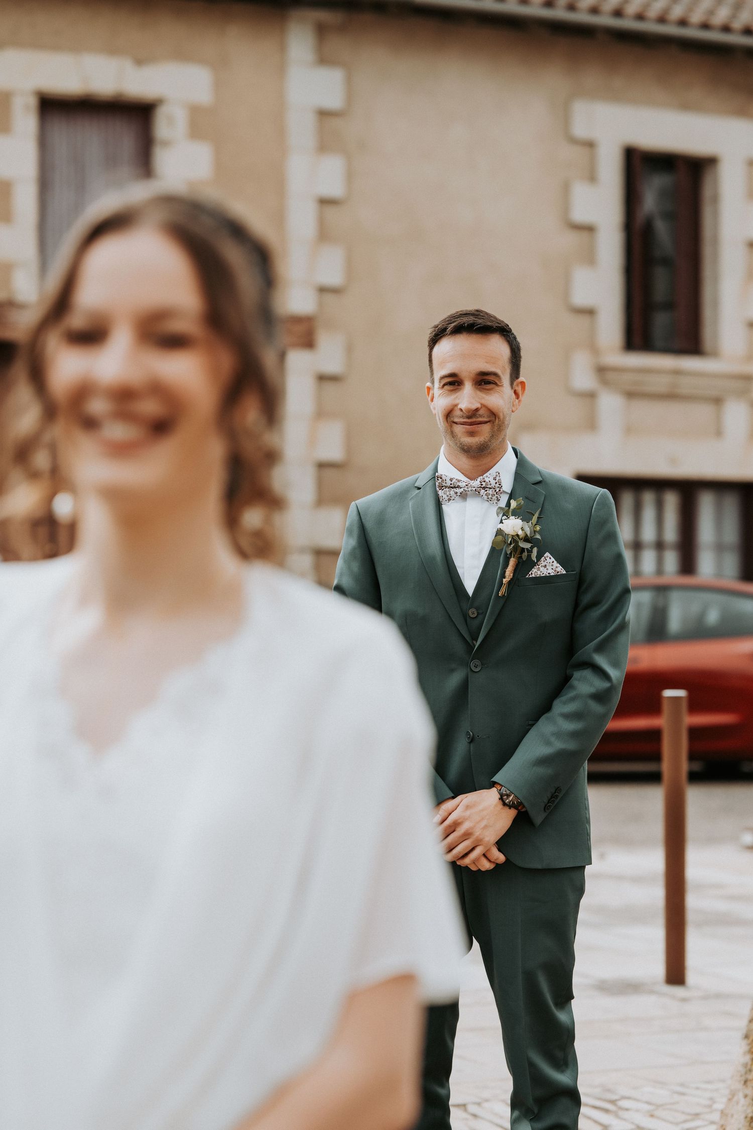 Groom in green suit smiles at blurred bride in white dress during outdoor wedding ceremony.