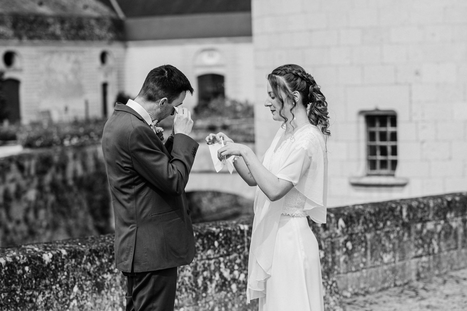 Emotional groom wiping tears as bride places wedding ring on his finger during outdoor ceremony.