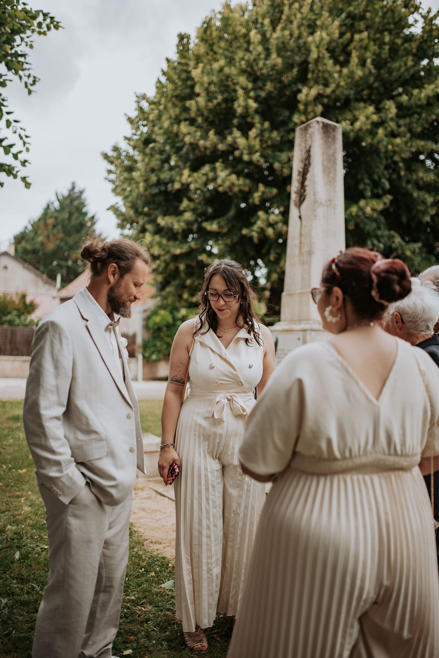 A group in white dresses and outfits gathers outdoors under leafy green trees in a candid wedding moment.