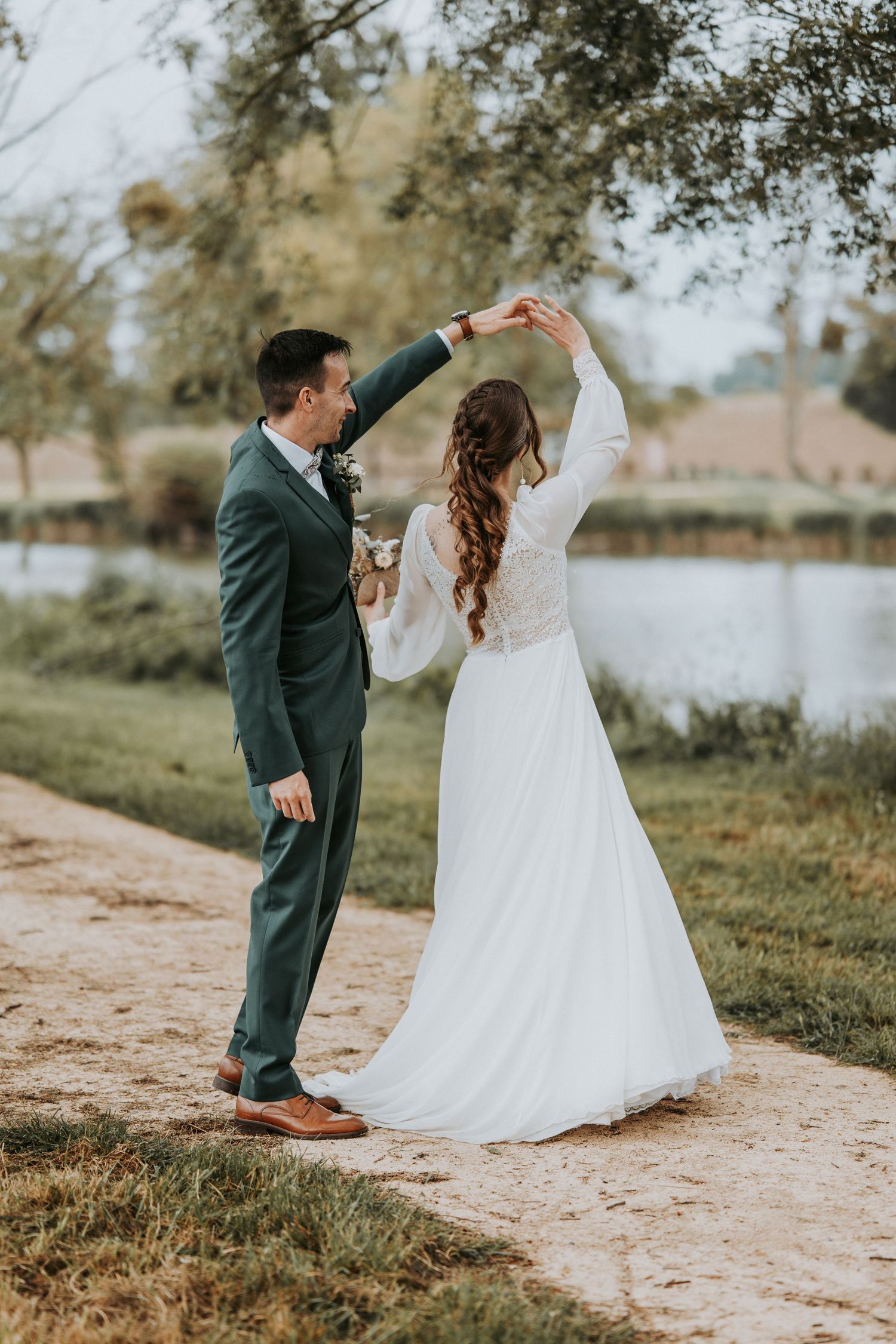 A groom in a green suit twirls his bride in a white lace wedding dress outdoors near a serene lake.