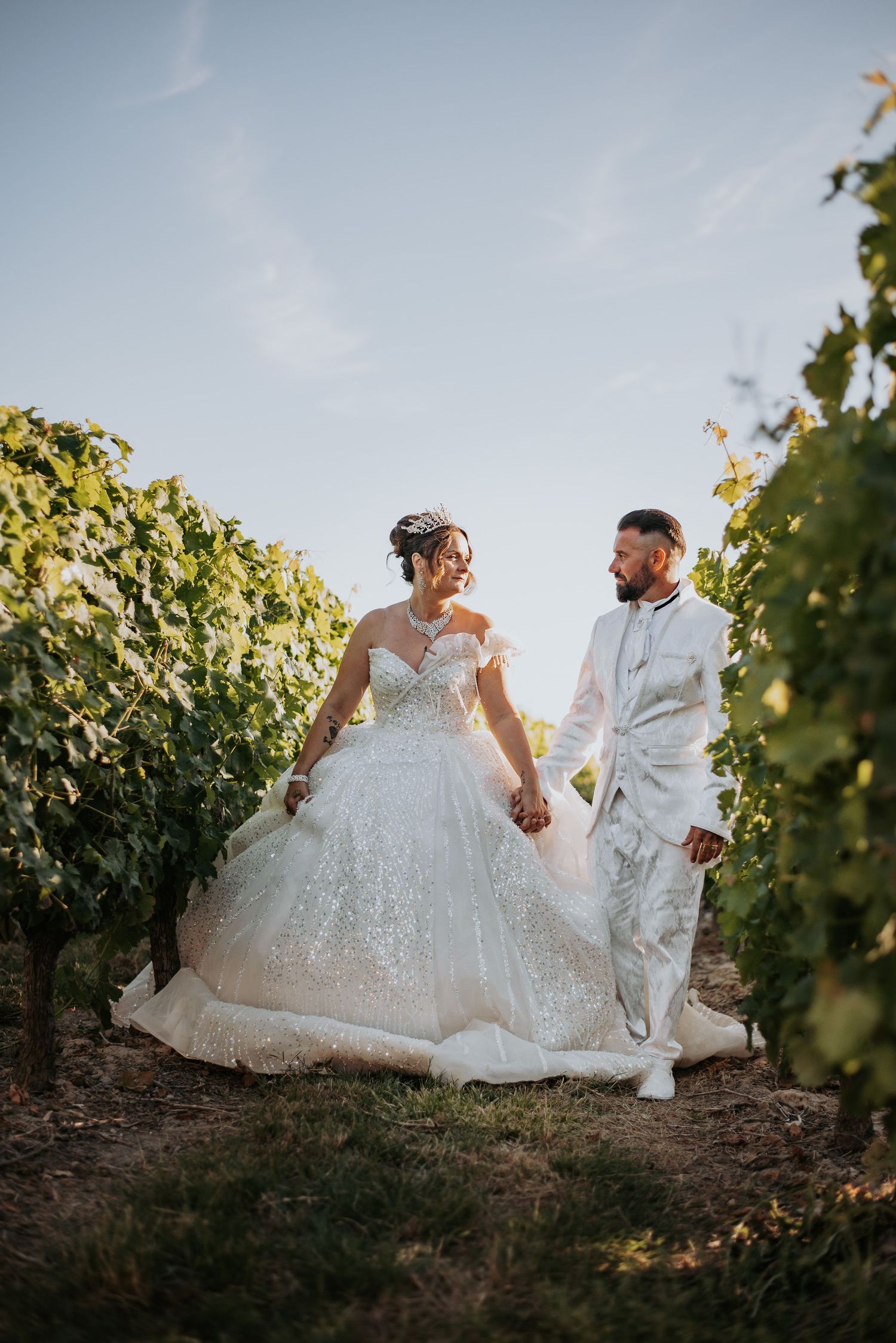 A couple in wedding attire walks together through a scenic vineyard at sunset.