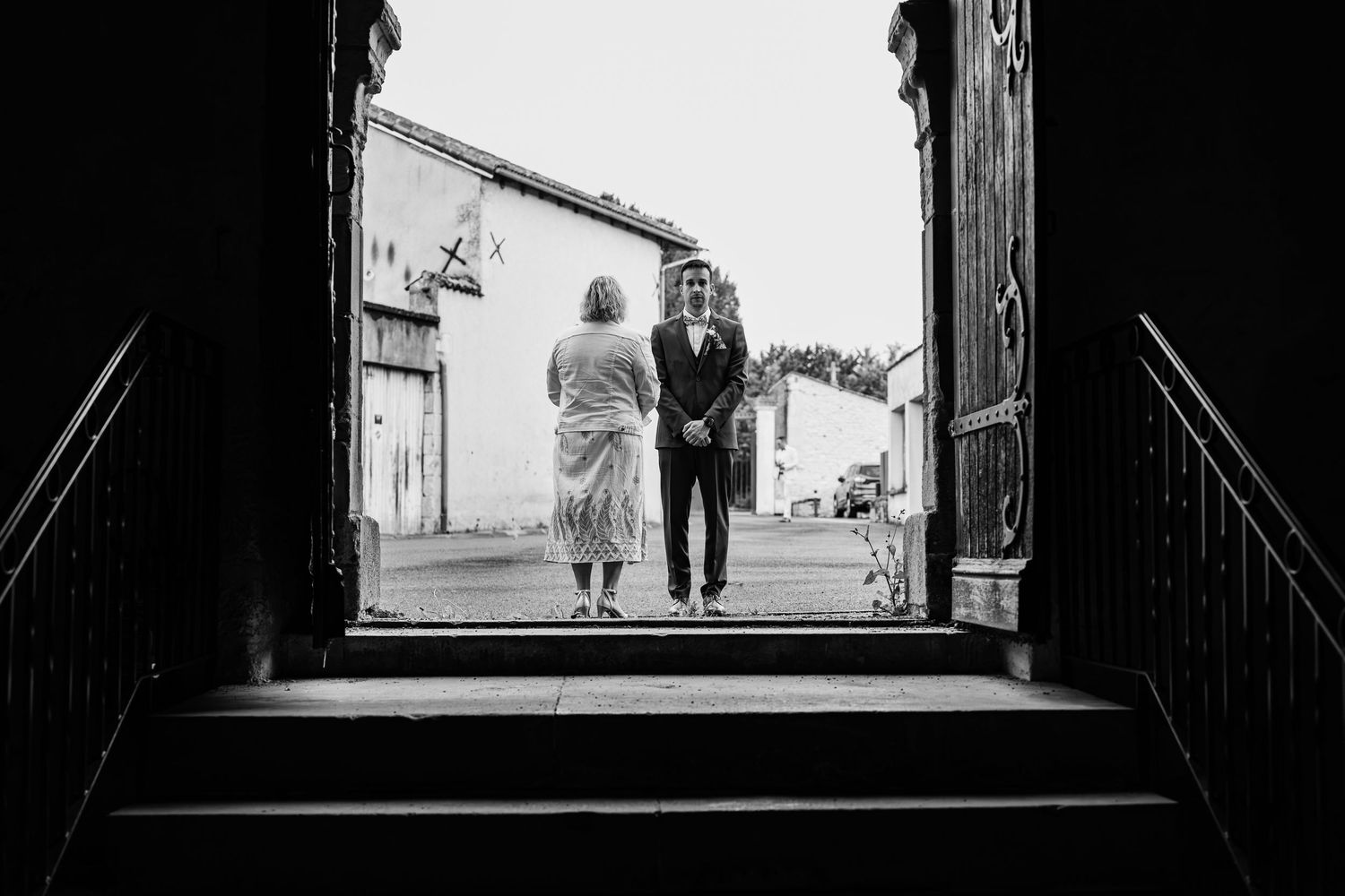 Black and white photo of a couple standing in a doorway framed by large wooden doors with steps leading outside.