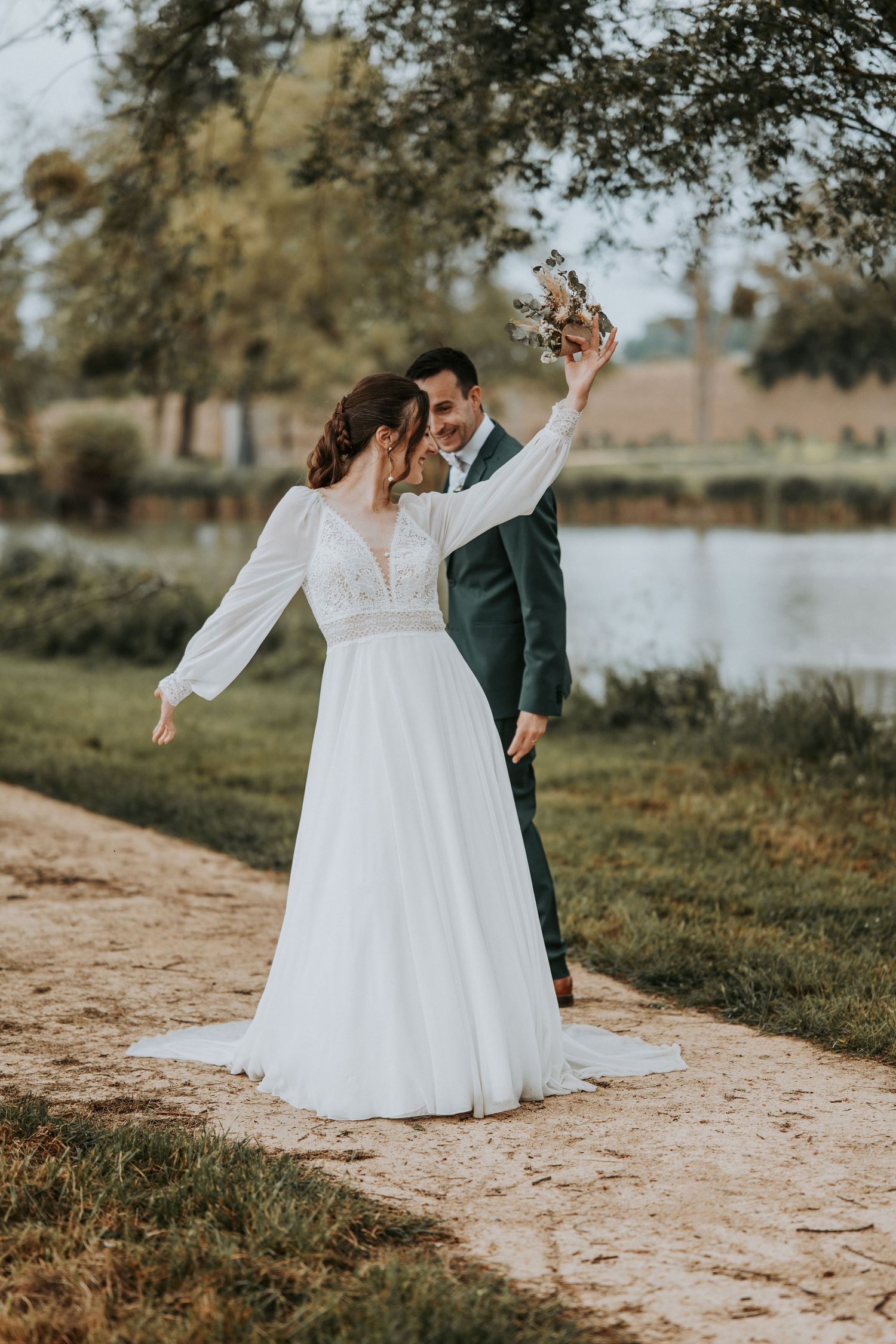 A bride in a white gown and groom in a dark suit celebrate outdoors near a pond, with the bouquet raised joyfully.