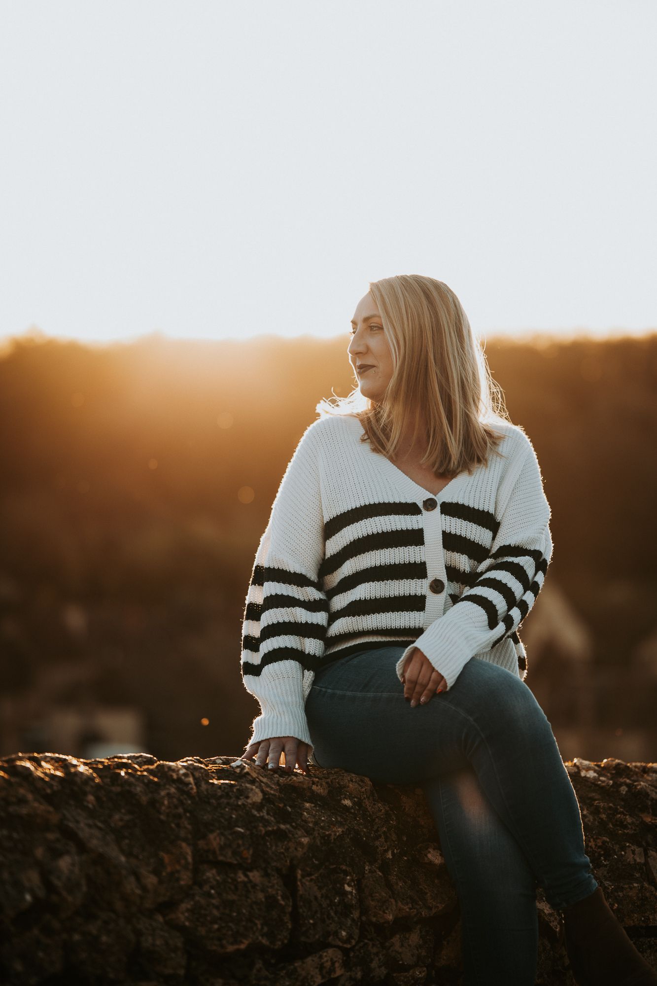 Woman in striped white sweater and jeans sits on rock wall at golden hour sunset.