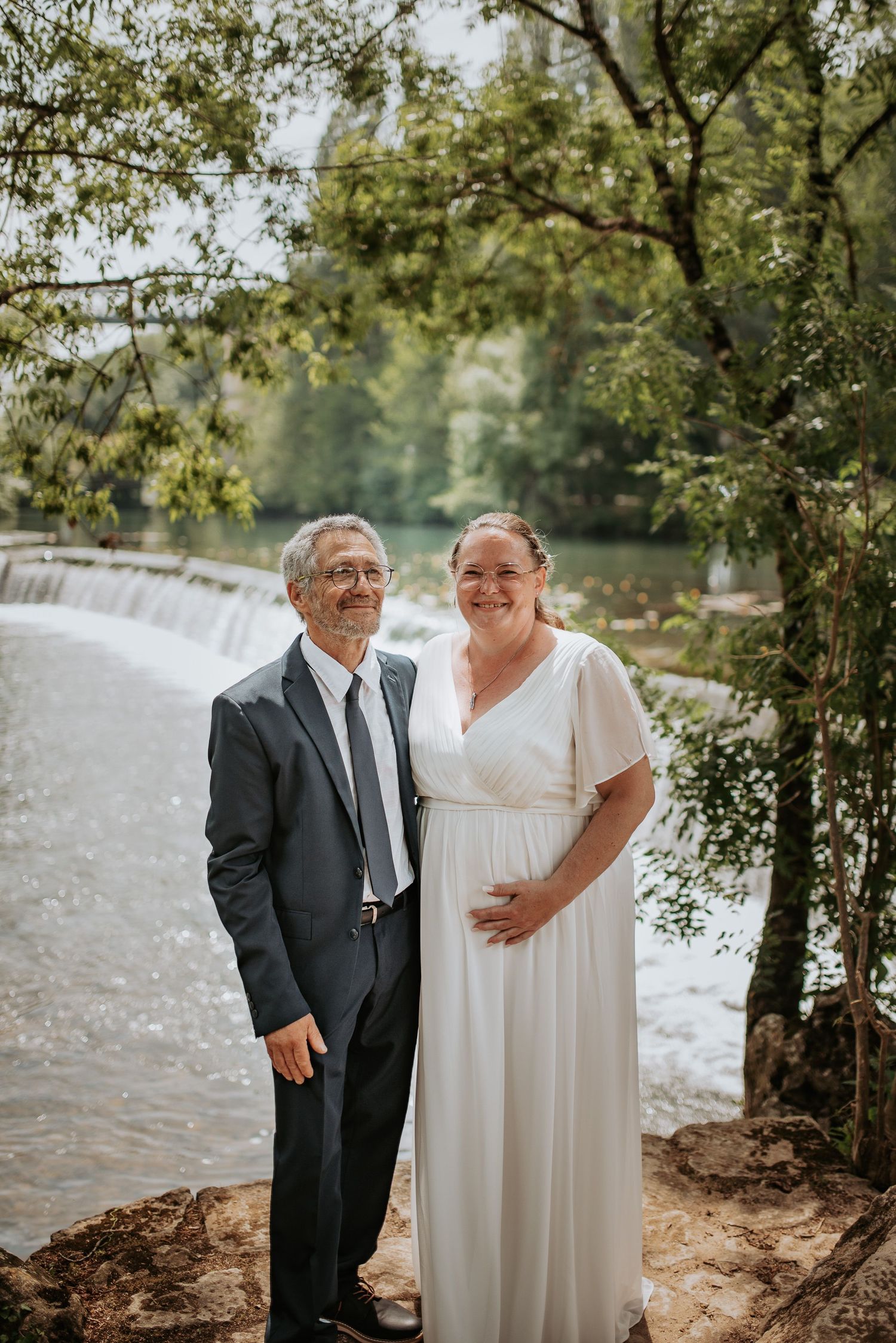 A couple in formal attire stands together by a scenic lakeside on a sunny day with trees in the background.