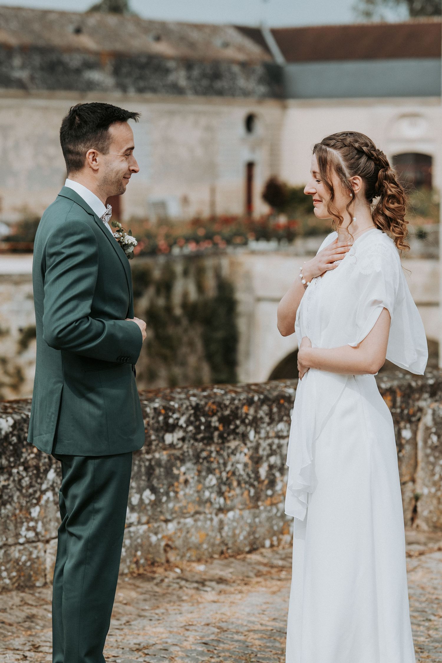A smiling bride in white dress and groom in green suit share a joyful moment outdoors at a rustic wedding venue.