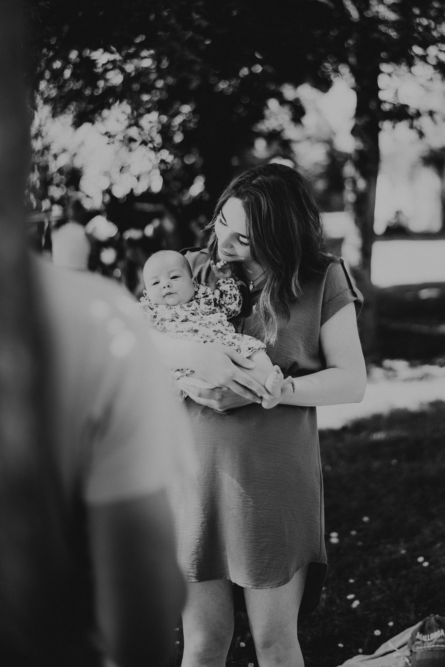 Black and white outdoor photo of a woman in a casual dress holding a small baby wrapped in a blanket during a sunny day.