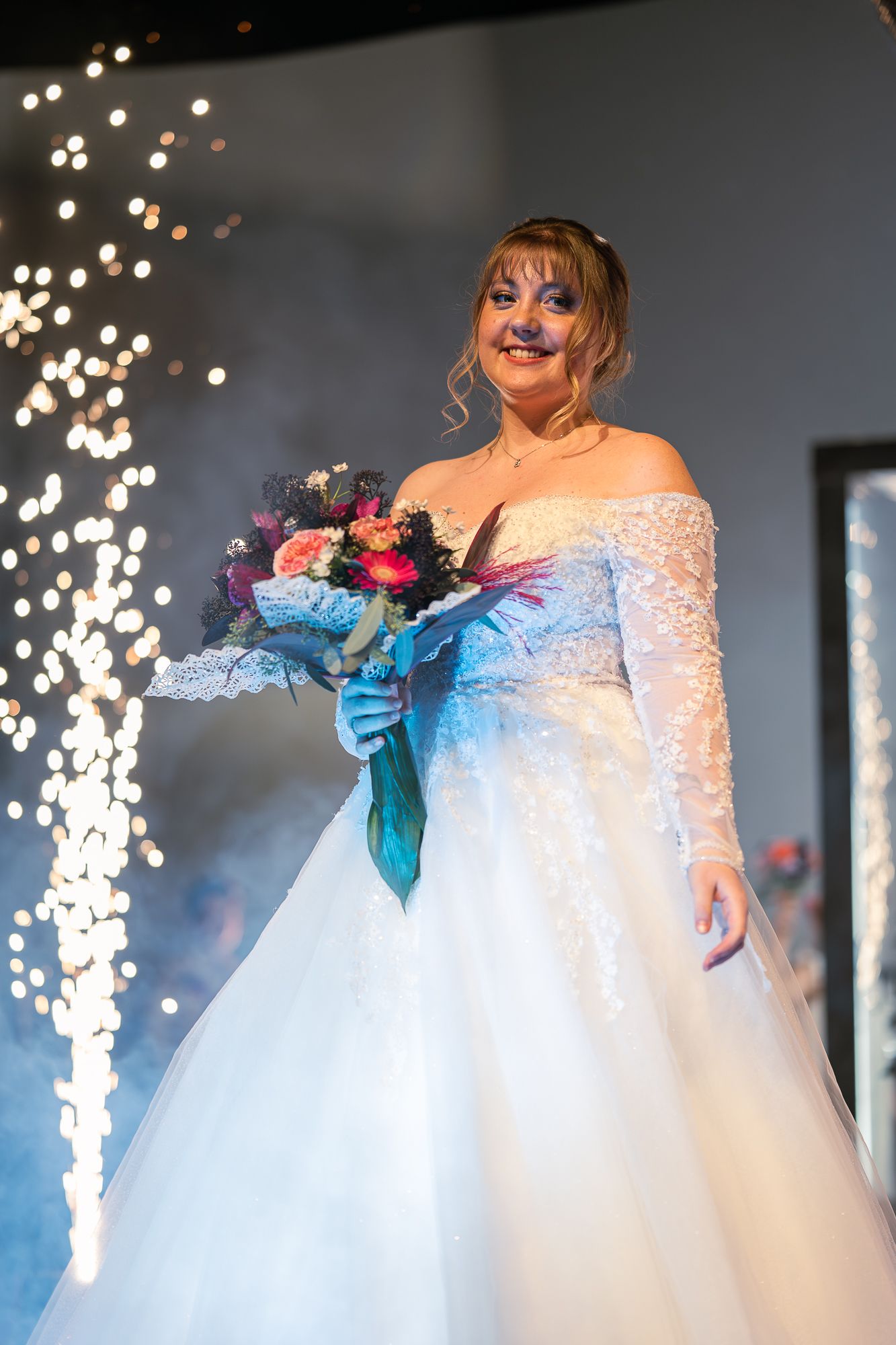A bridal fashion show featuring a flowing white ballgown with lace sleeves and sparkling pyrotechnics in the background.