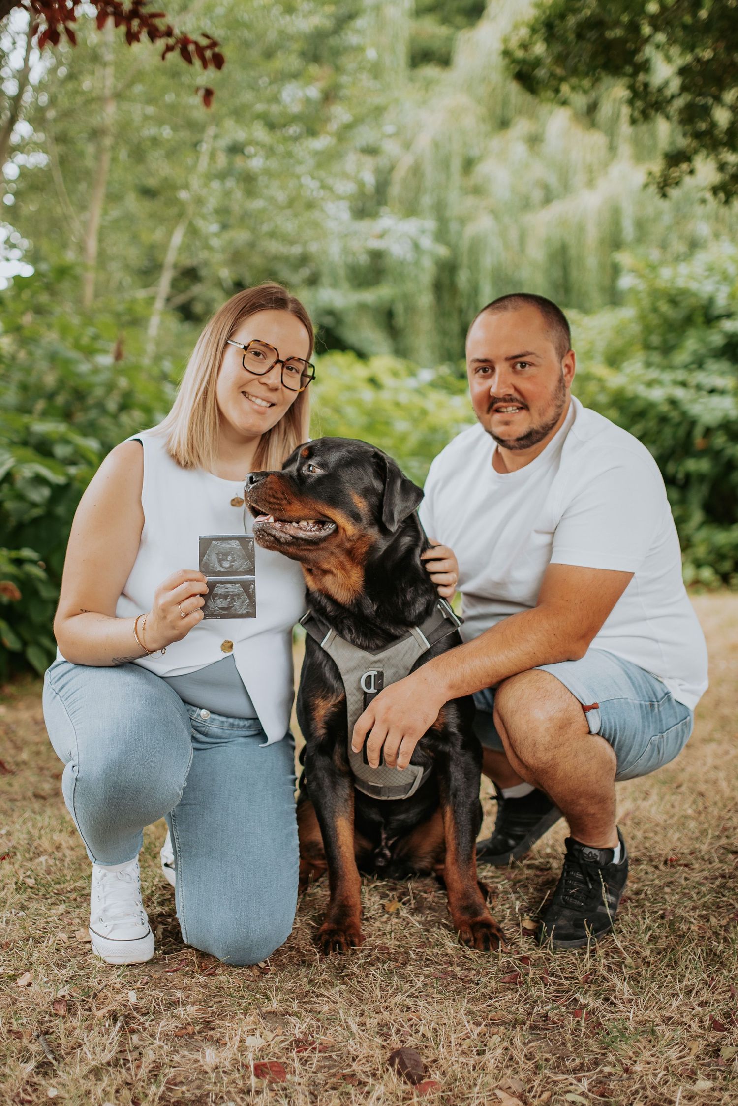 A Rottweiler pet dog poses outdoors with two people in a natural woodland setting.