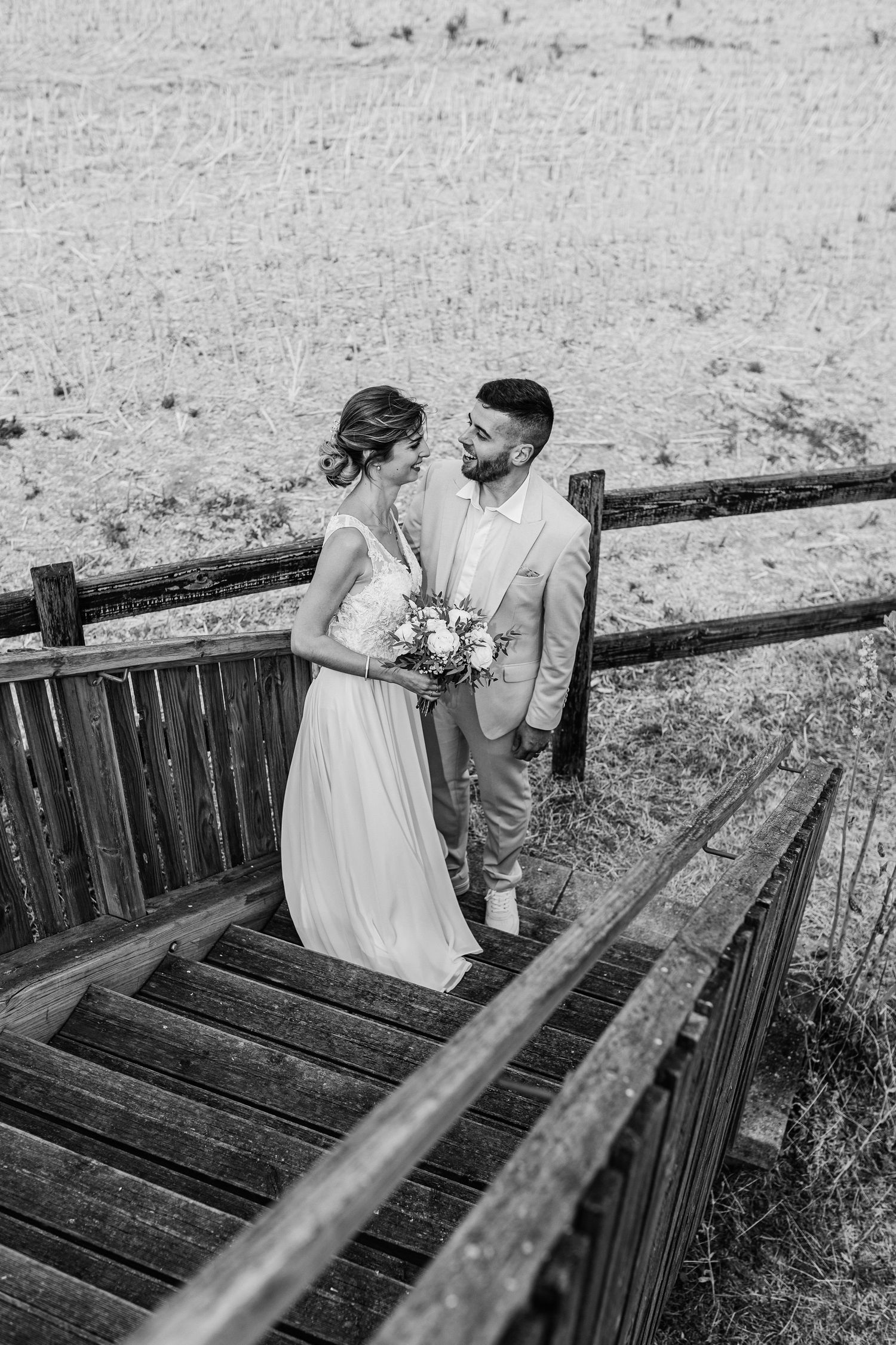 Black and white wedding photo of a couple standing on rustic wooden steps with a bouquet and rustic fence in background.
