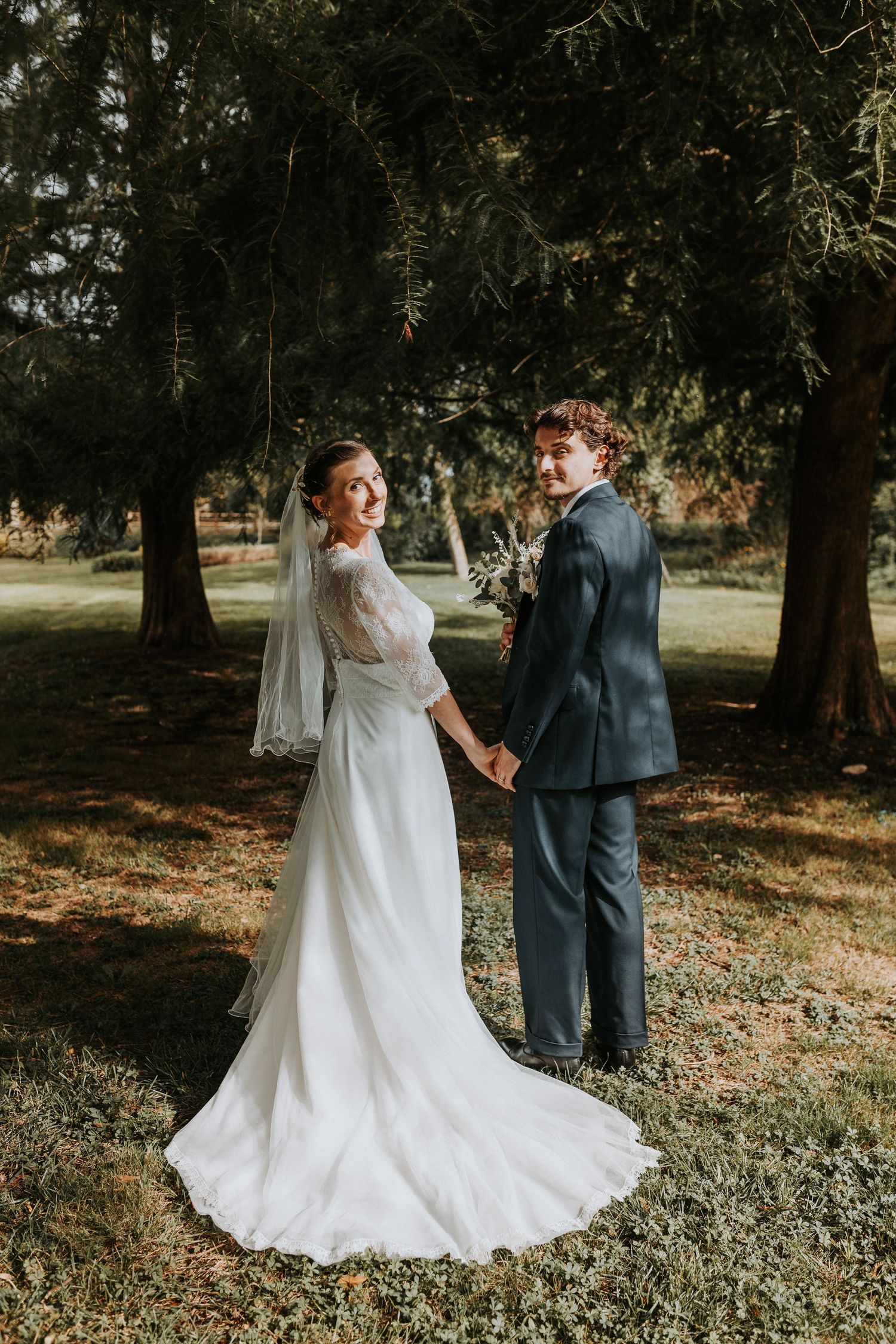 A couple in wedding attire holds hands while standing in a wooded outdoor setting on their special day.