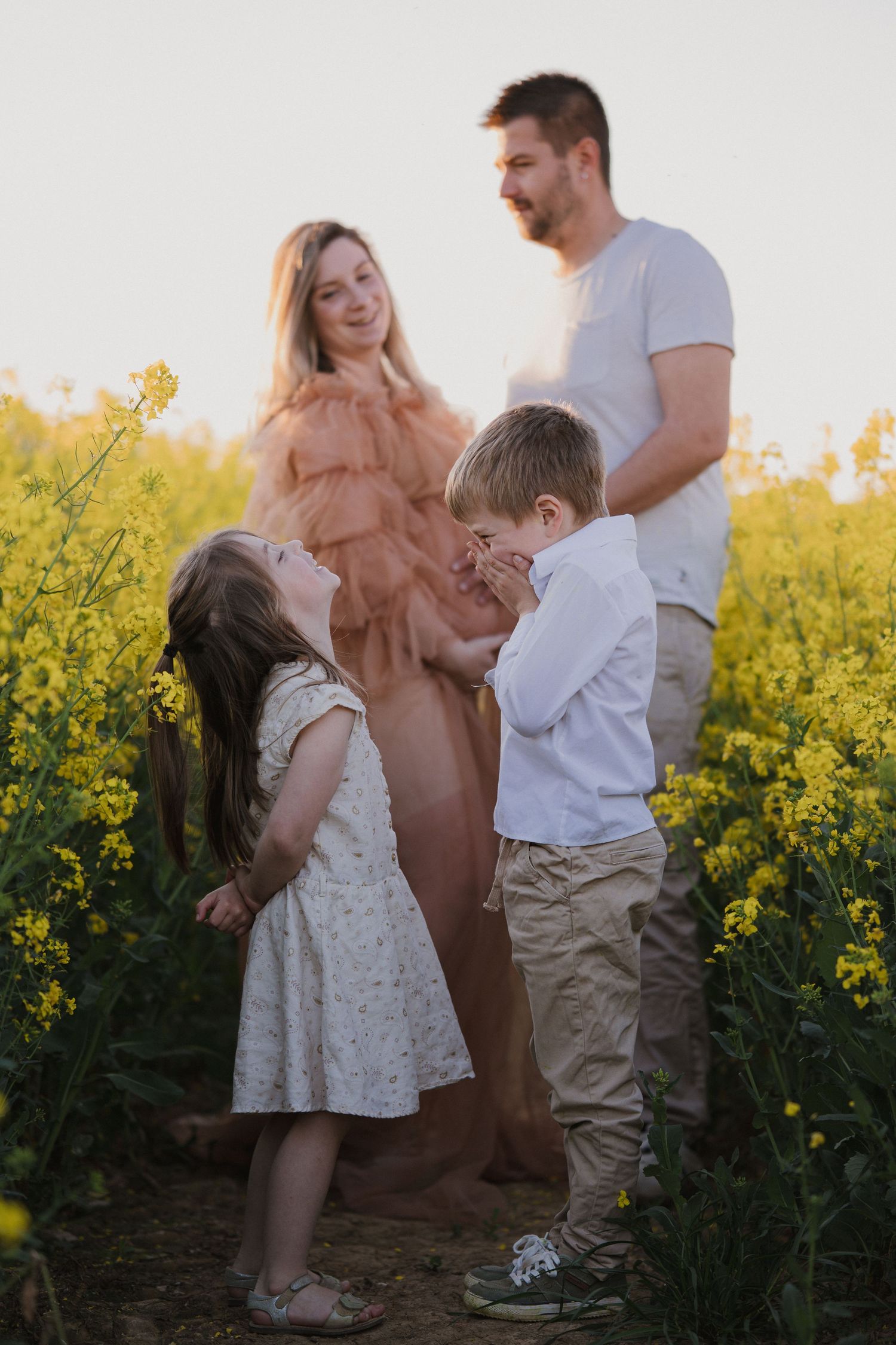Family of four with pregnant mother in peach dress standing in yellow flower field at golden hour sunset.