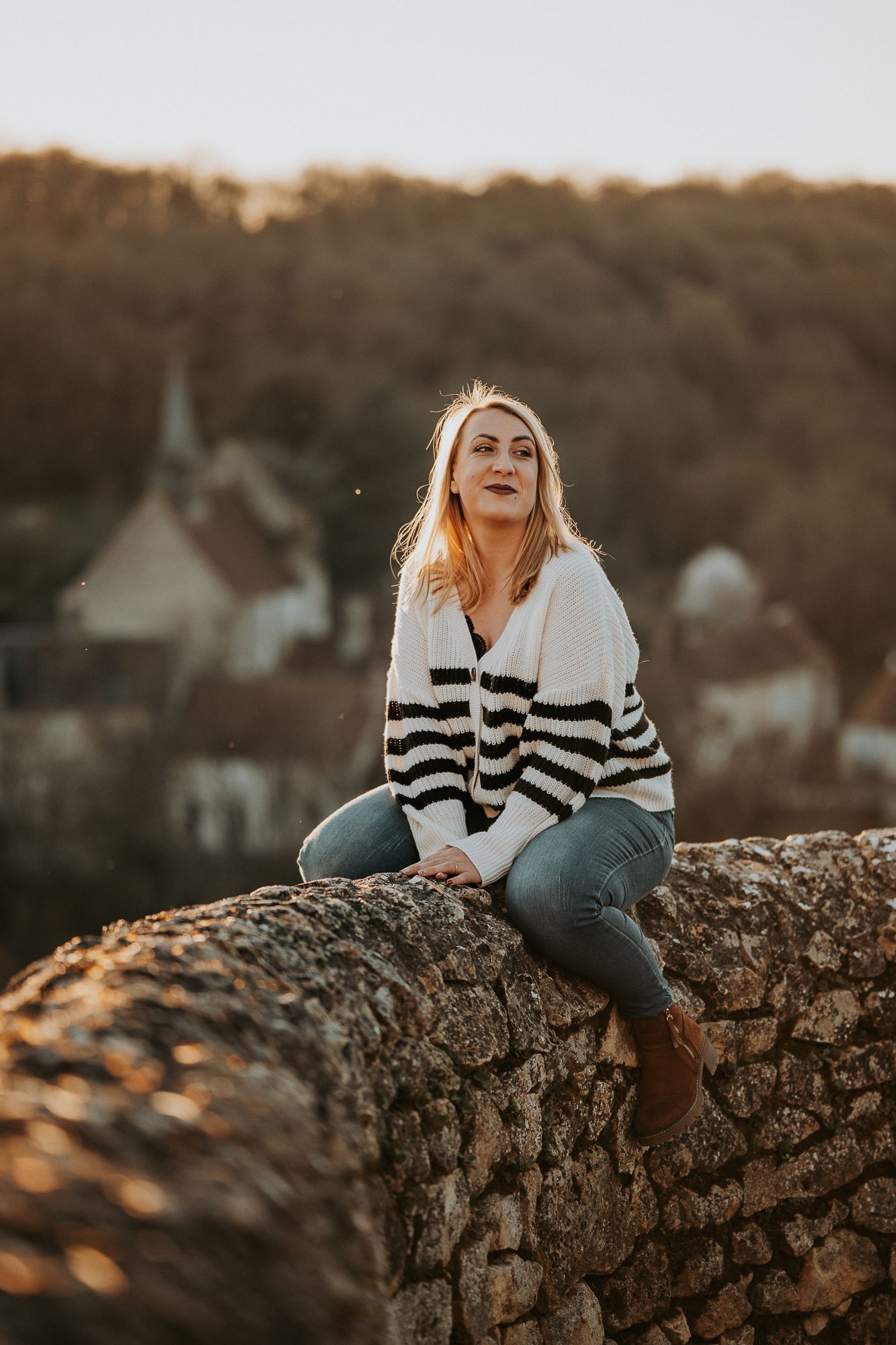 A blonde woman sits on a stone wall in a striped cardigan and jeans, with a golden-hour village backdrop.