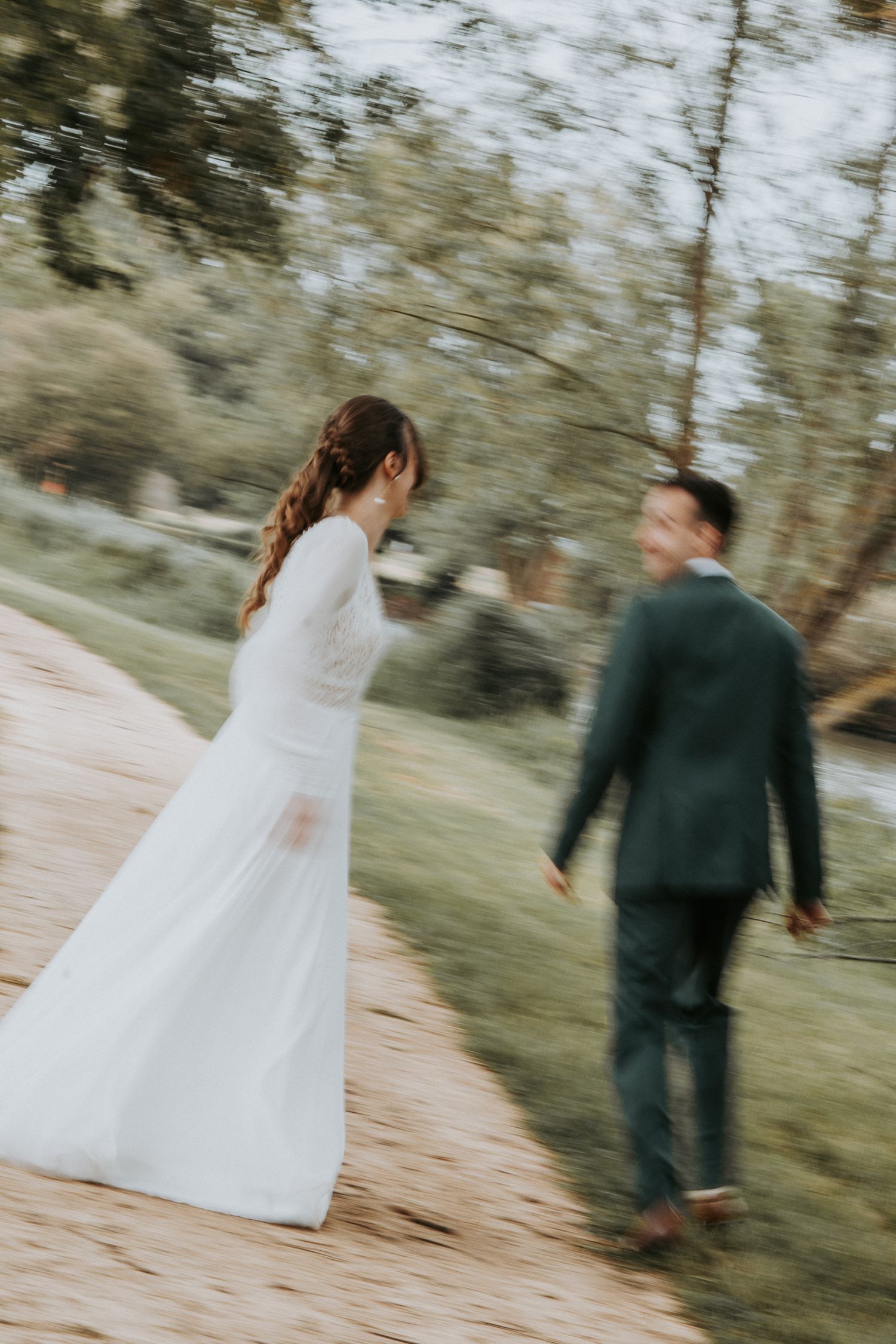 Bride in white gown and groom in dark suit walk together on a path outdoors in a blurred motion wedding photo.