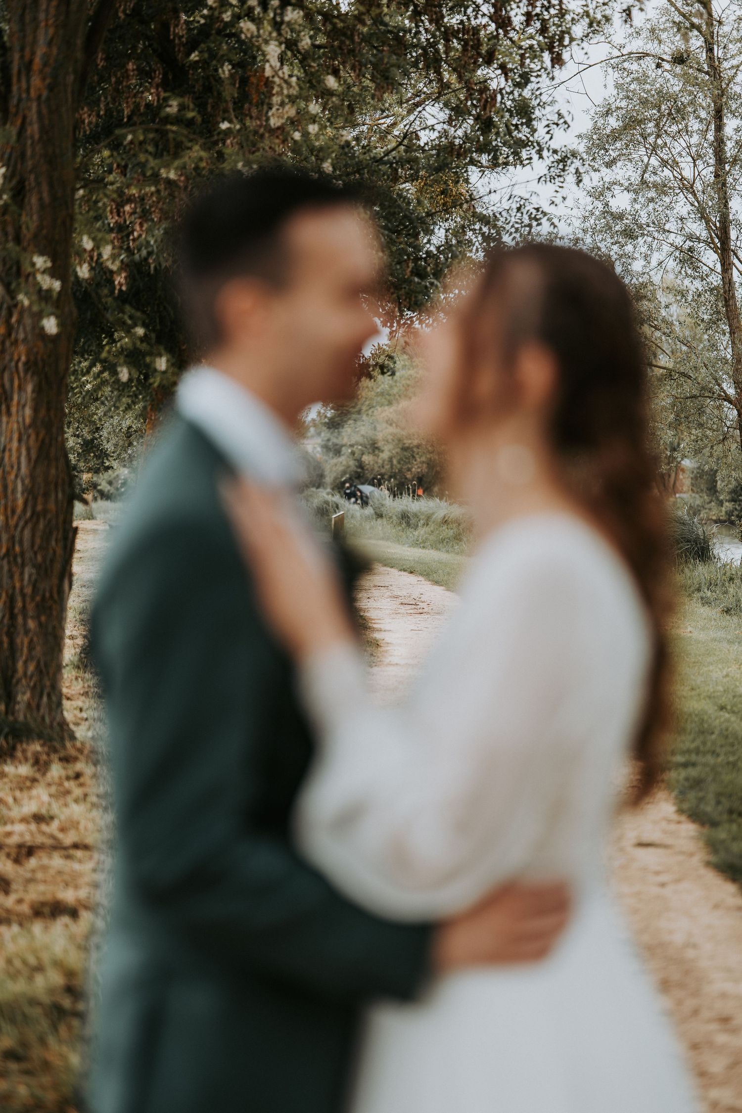 Blurred wedding couple embracing outdoors among trees, bride in white gown and groom in dark suit.