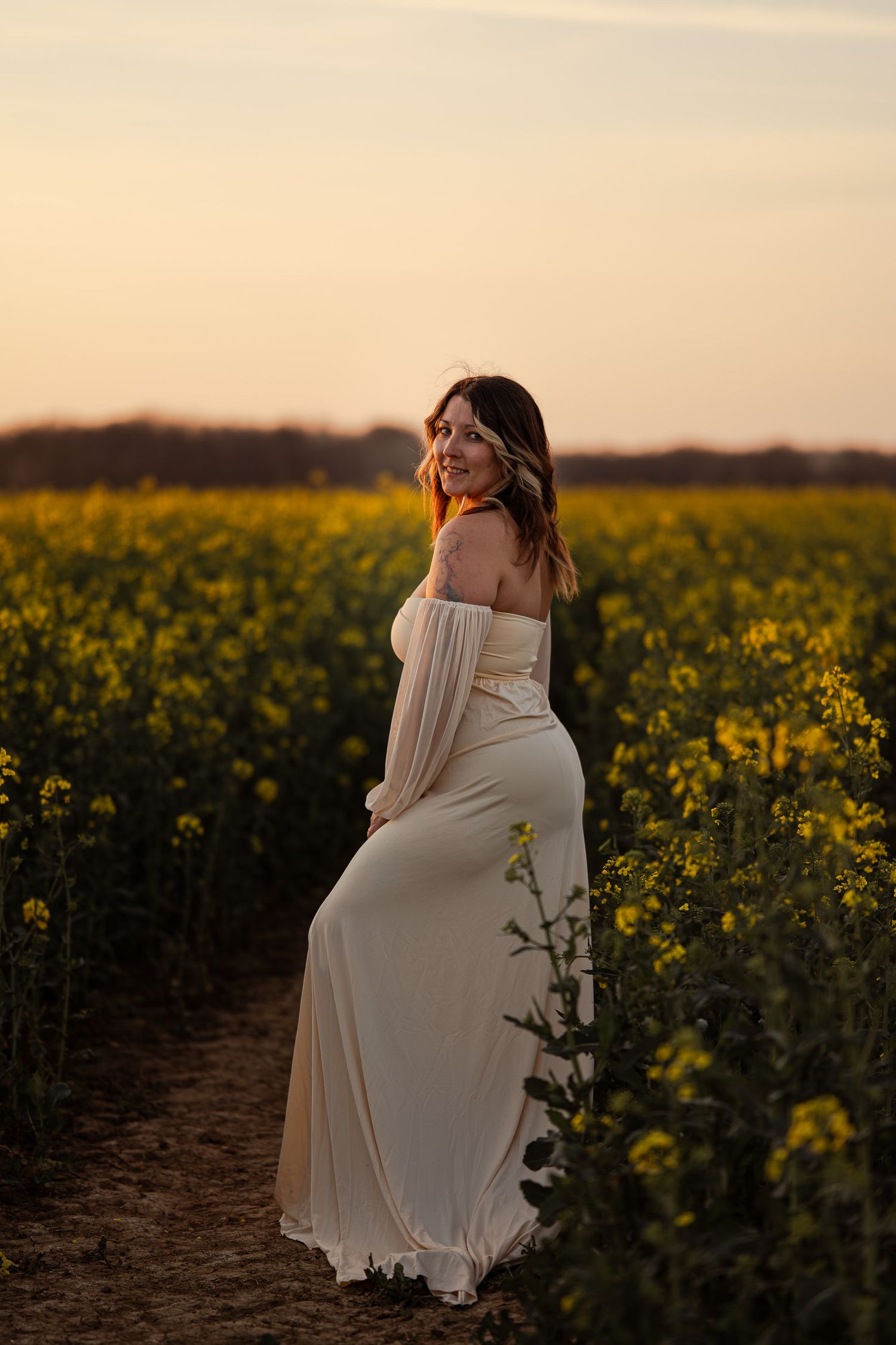Woman in white off-shoulder gown stands in yellow flower field at golden sunset.