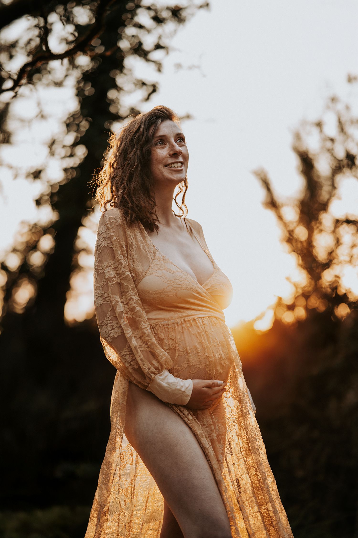 Pregnant woman in golden lace gown glowing in sunset light, smiling serenely outdoors.