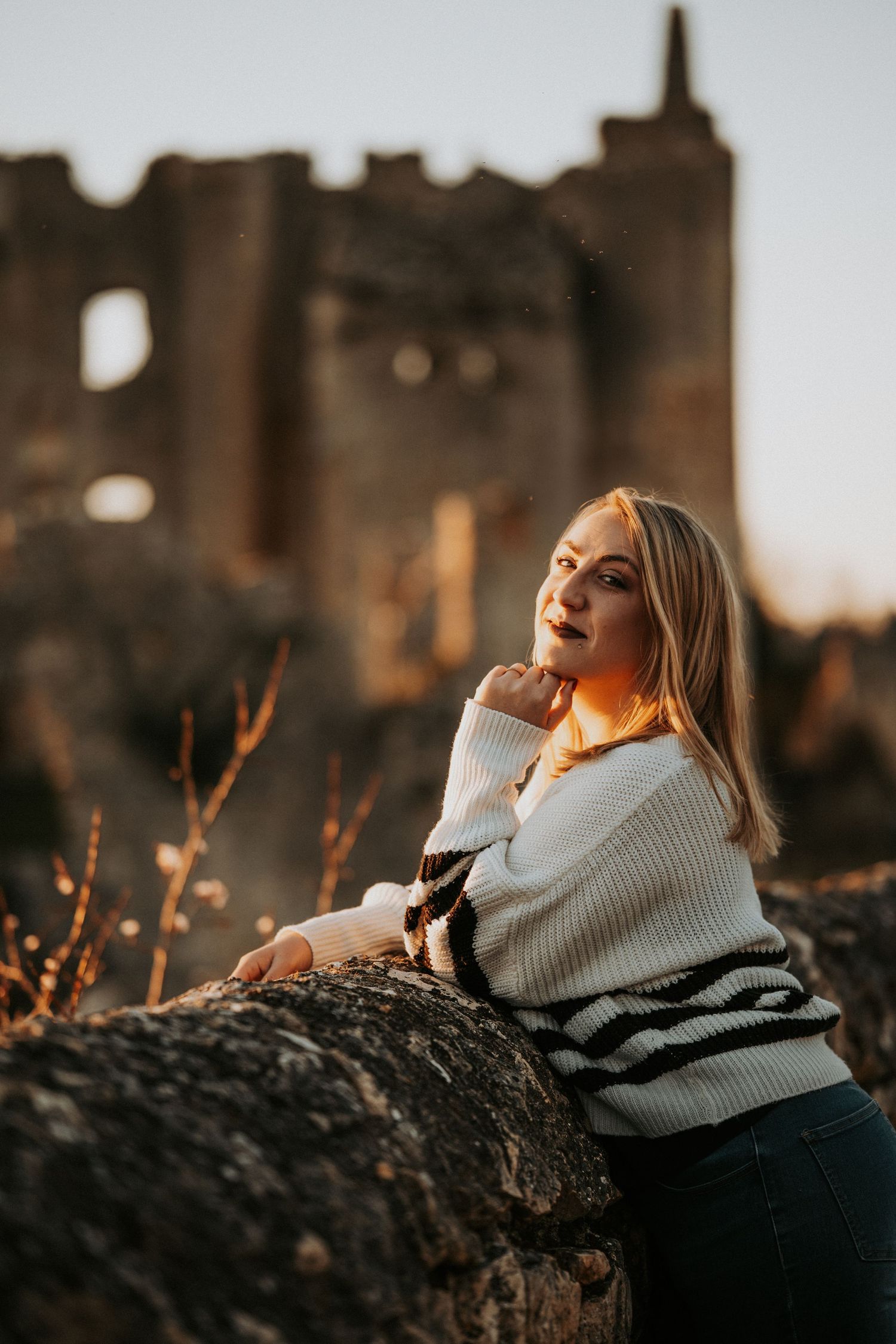 Blonde woman in white sweater leaning on stone wall at golden hour with ancient castle ruins in background.