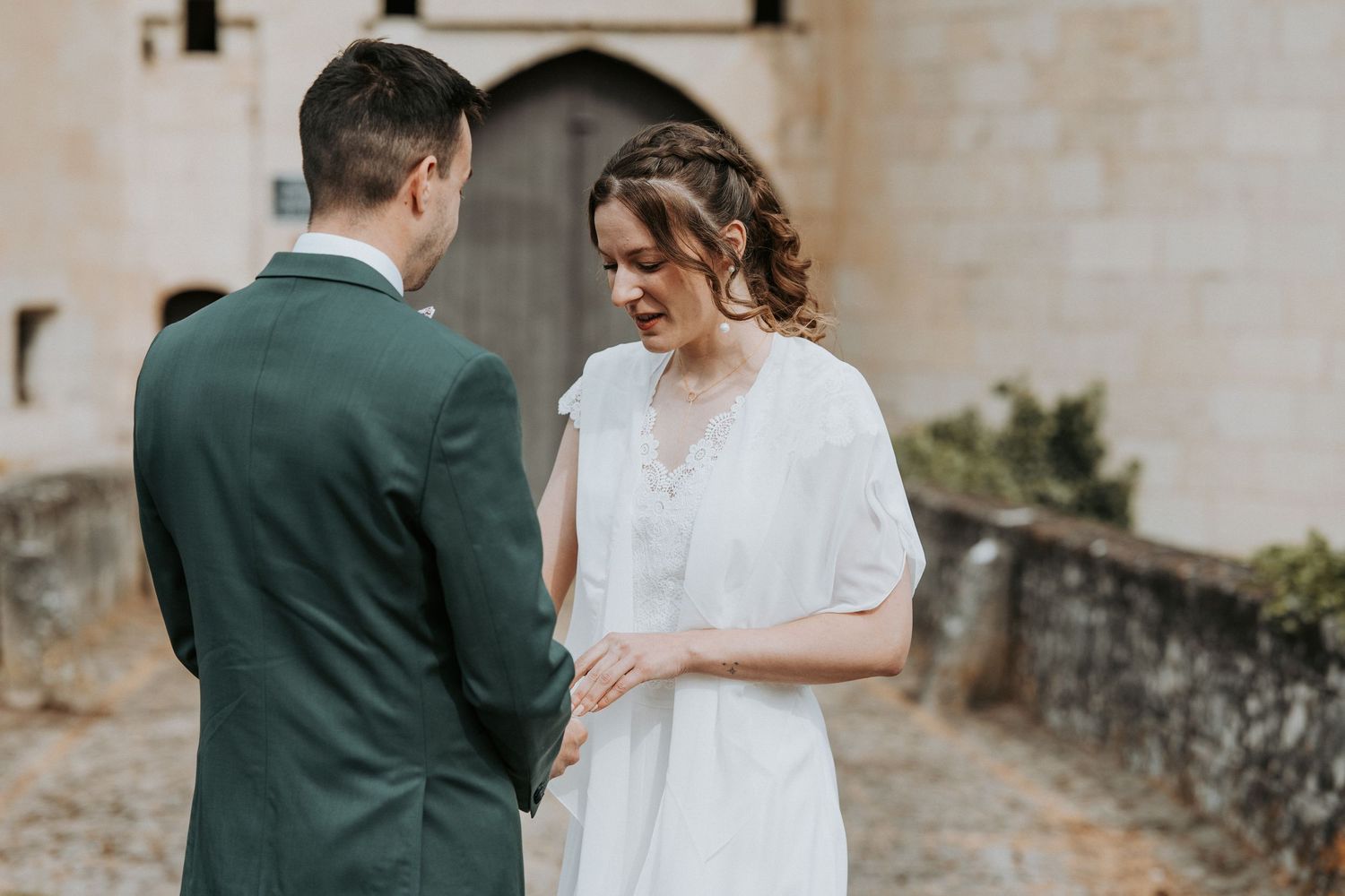 A bride in a white dress and groom in a green suit hold hands during an outdoor wedding ceremony by a stone wall.