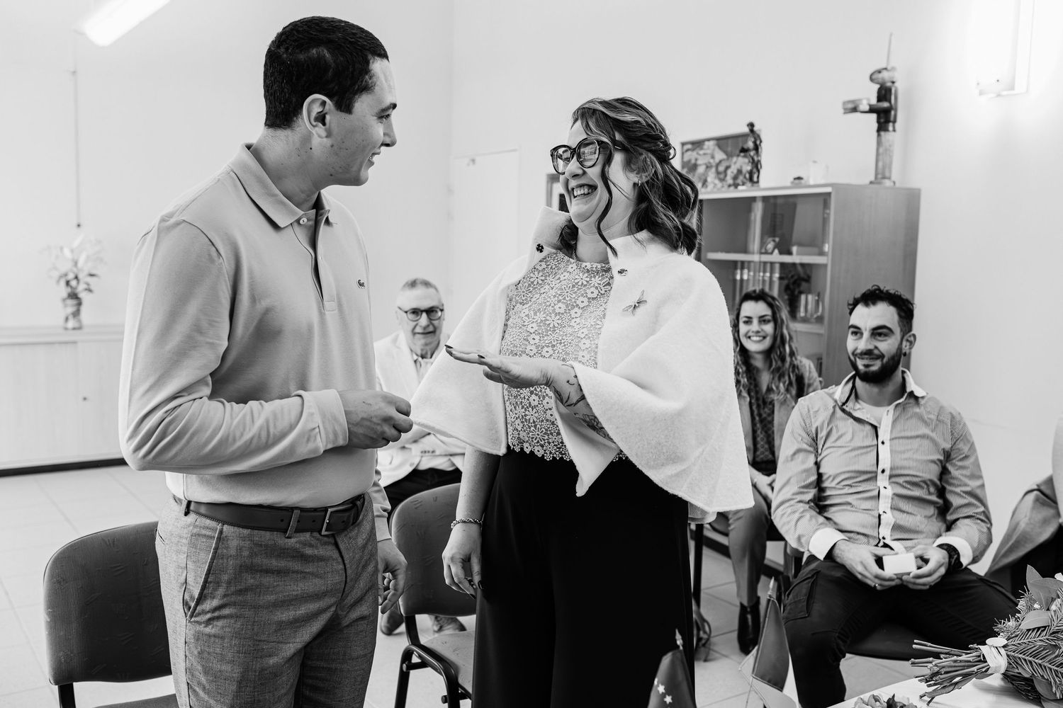 Man and woman smiling and exchanging gifts at an indoor gathering with guests watching in background.