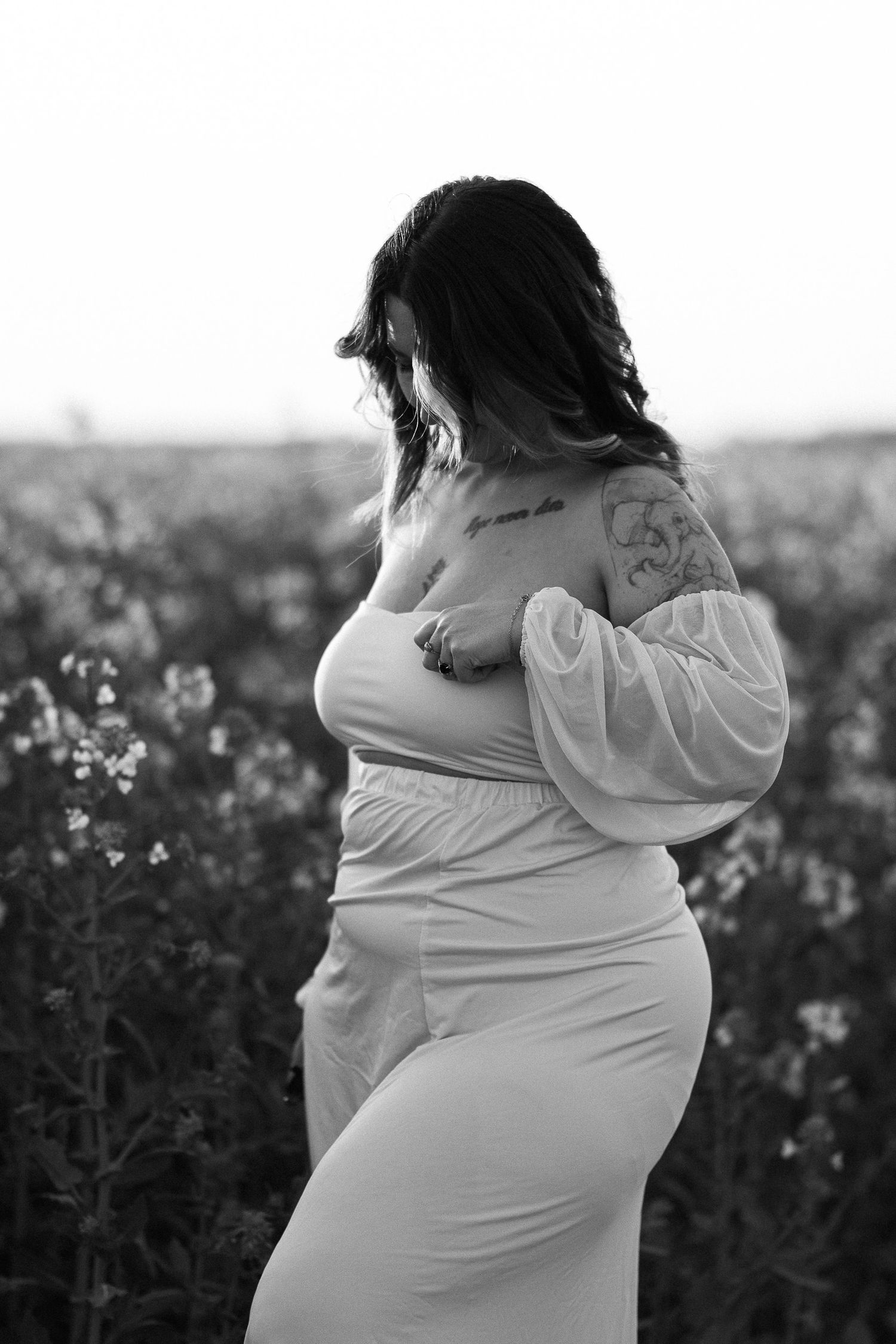 Black and white photo of a pregnant woman with tattoos in a flowy white dress standing in a blooming flower field.