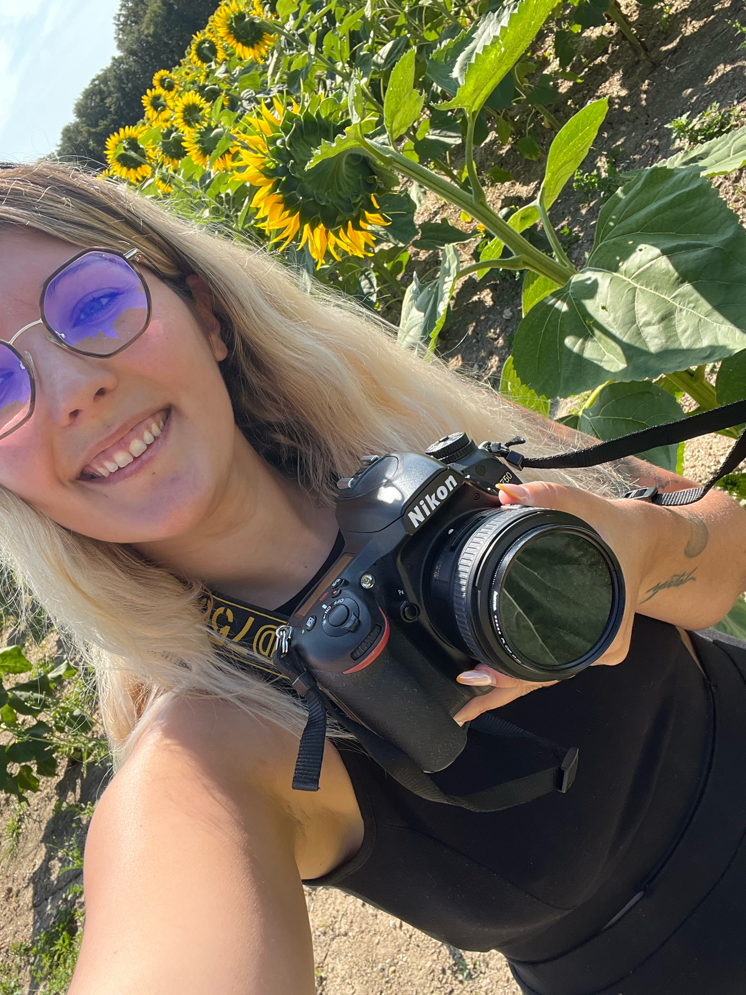 Someone holding a Nikon camera takes a sunny selfie near bright yellow sunflowers wearing purple tinted glasses.