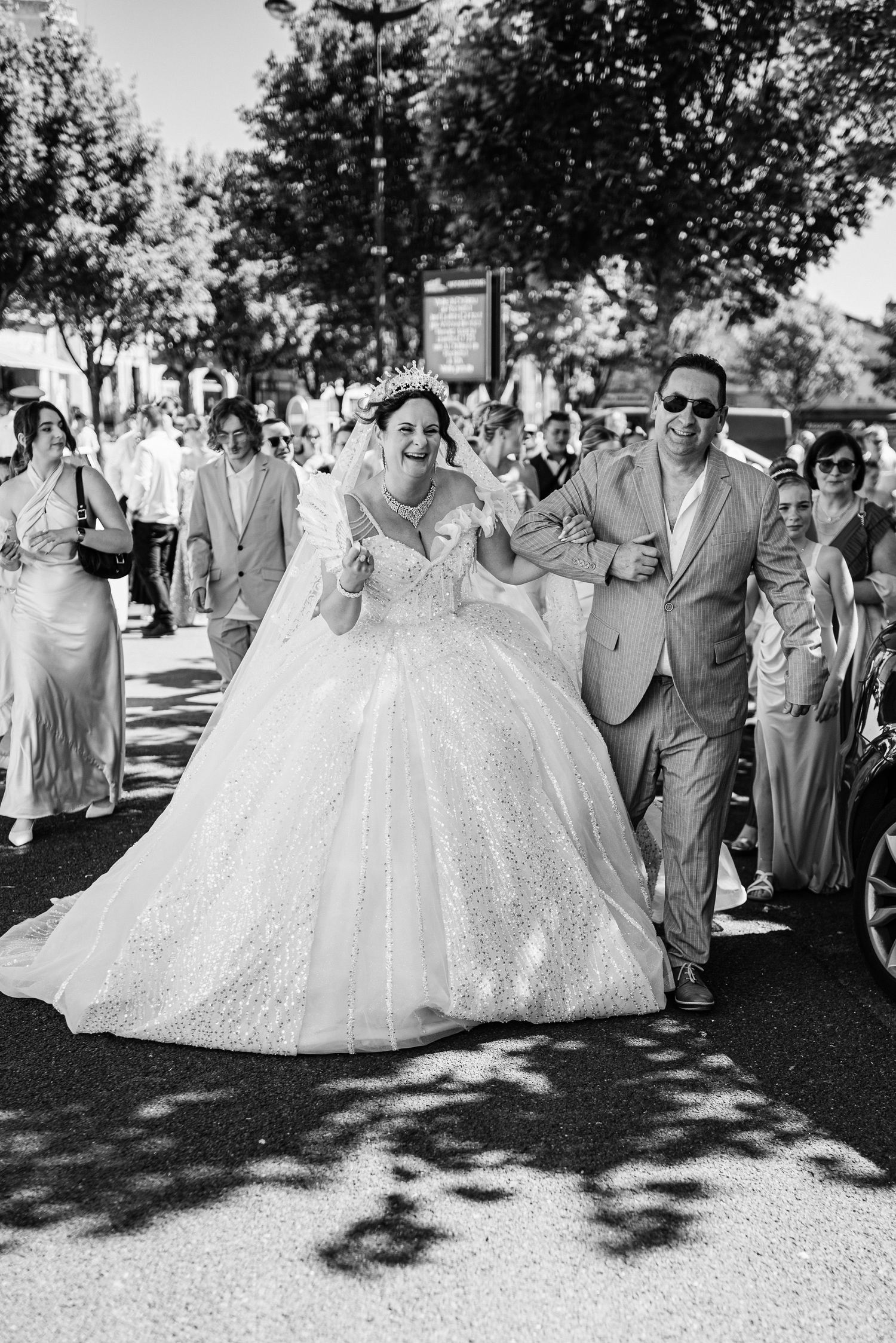 A wedding party walks down a tree-lined street with the bride wearing a stunning white ball gown and guests following behind.