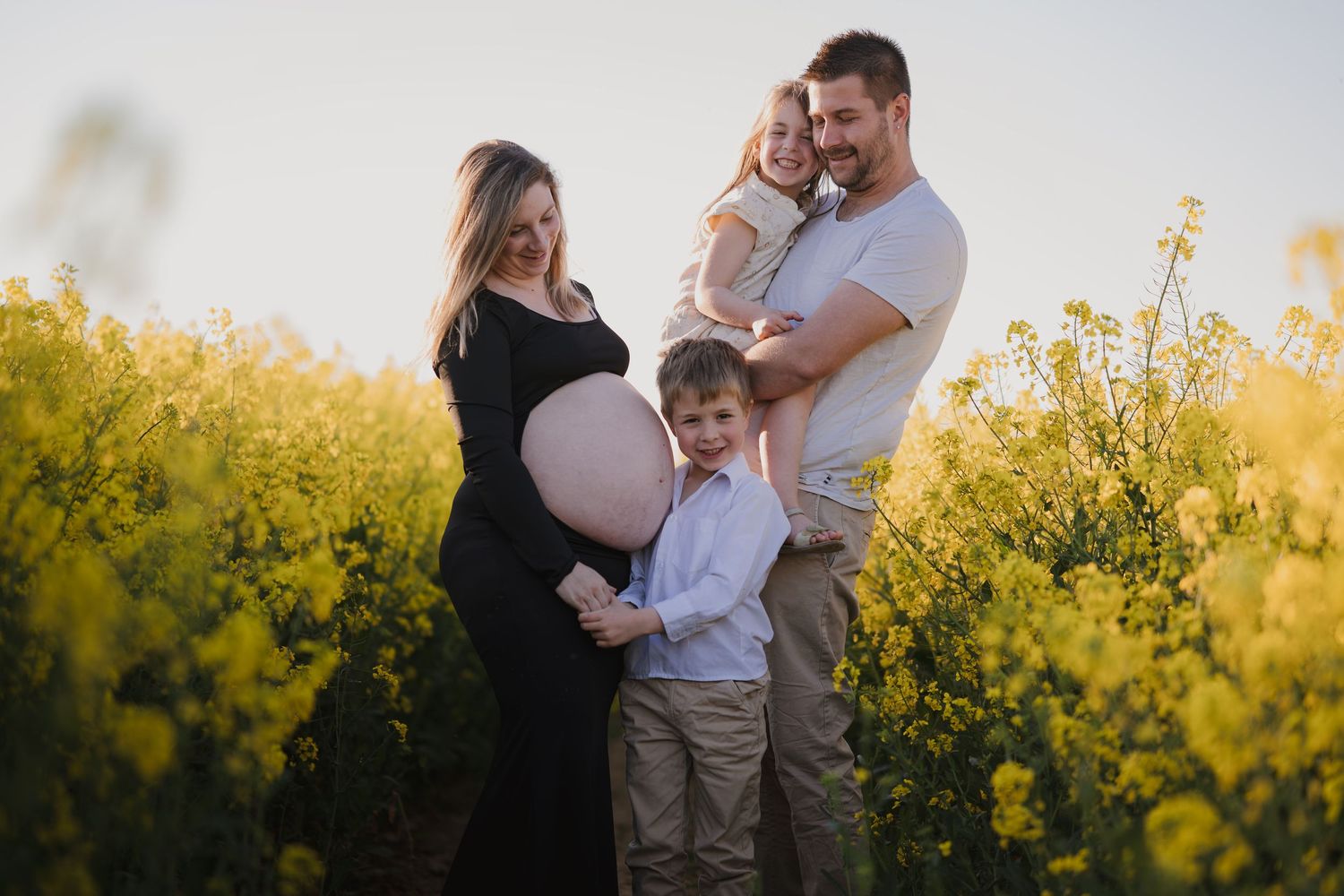 Pregnant woman in black dress with family in yellow flower field during golden hour maternity photoshoot.