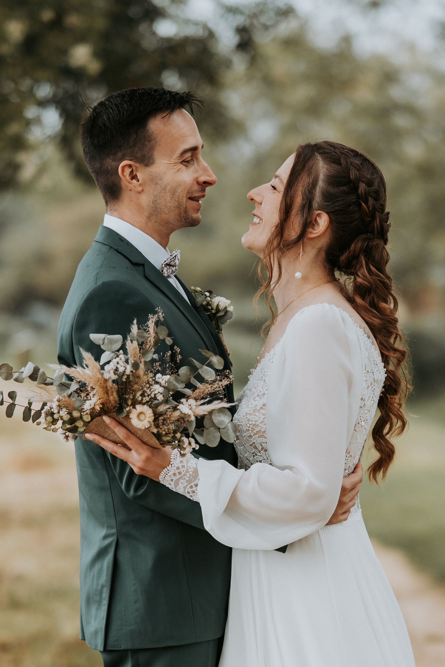 A bride and groom share a joyful moment, holding a dried floral bouquet outdoors on their wedding day.