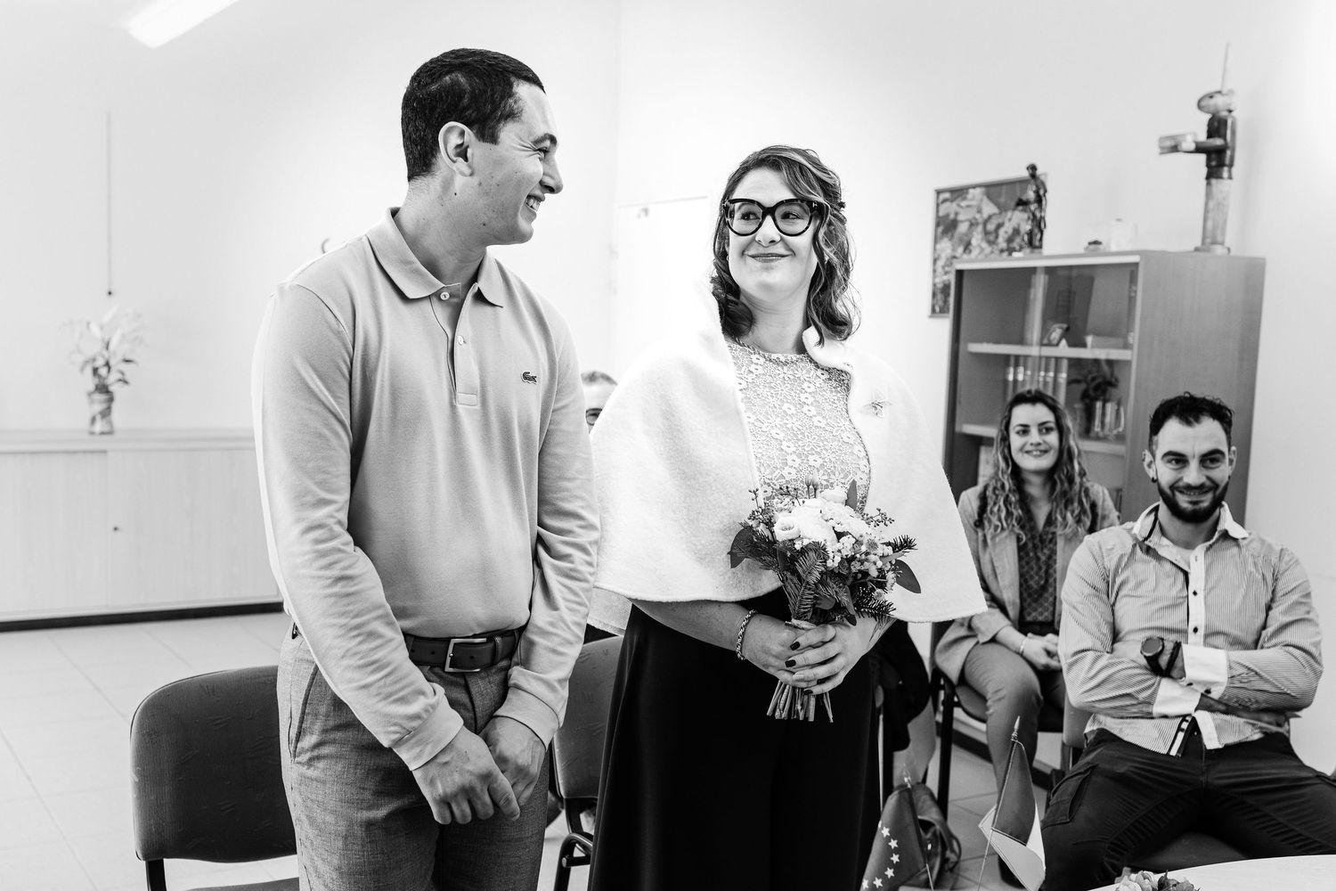 Black and white photo of a smiling couple holding flowers, with guests seated in background at a casual indoor celebration.