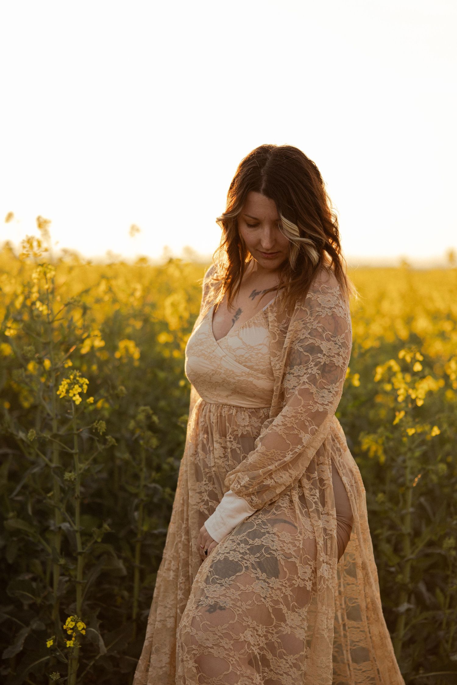 Woman in lace dress standing in golden rapeseed field at sunset, looking down with soft natural light.