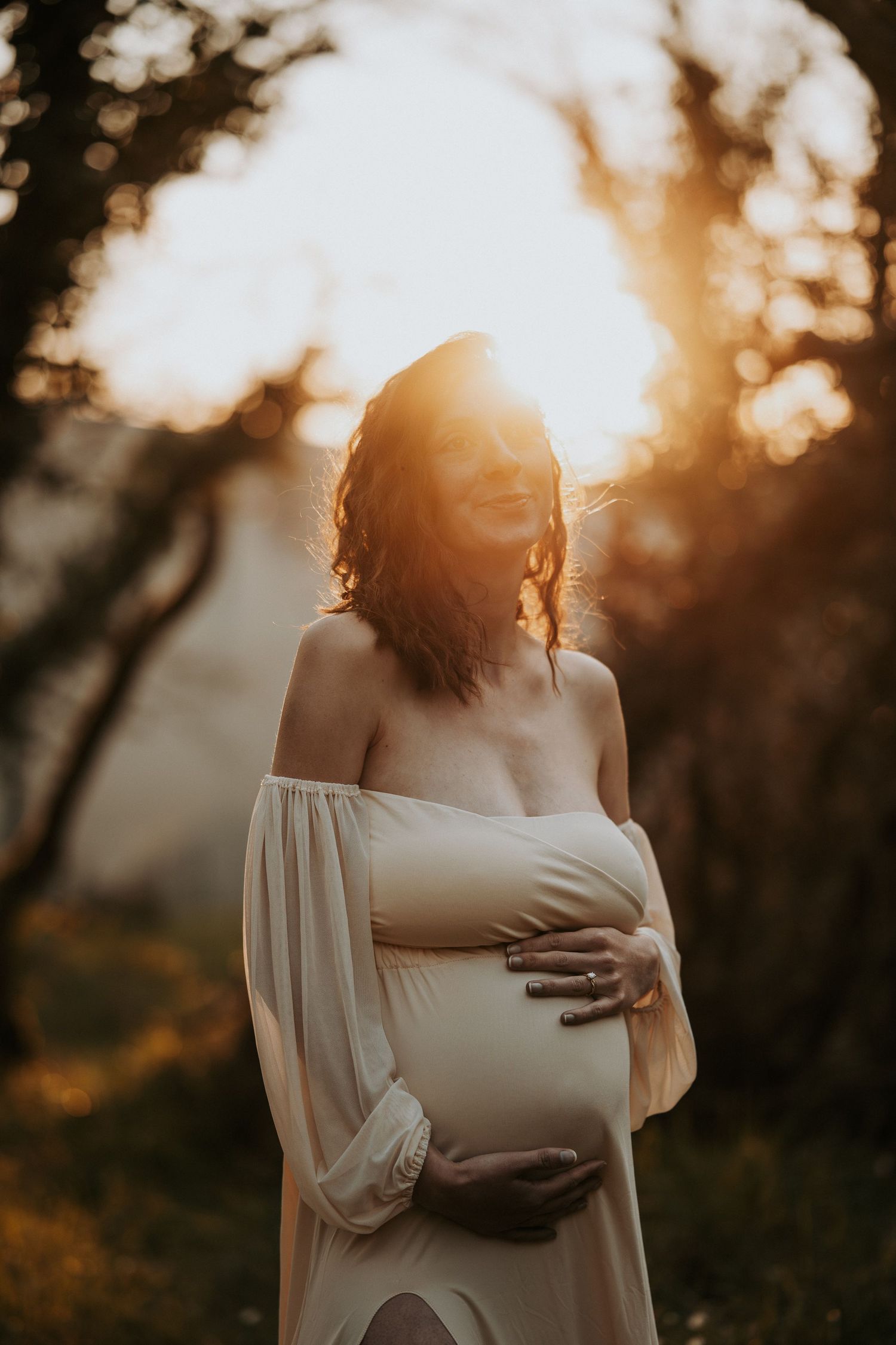 Pregnant woman in flowing off-shoulder gown cradling baby bump outdoors at golden sunset.