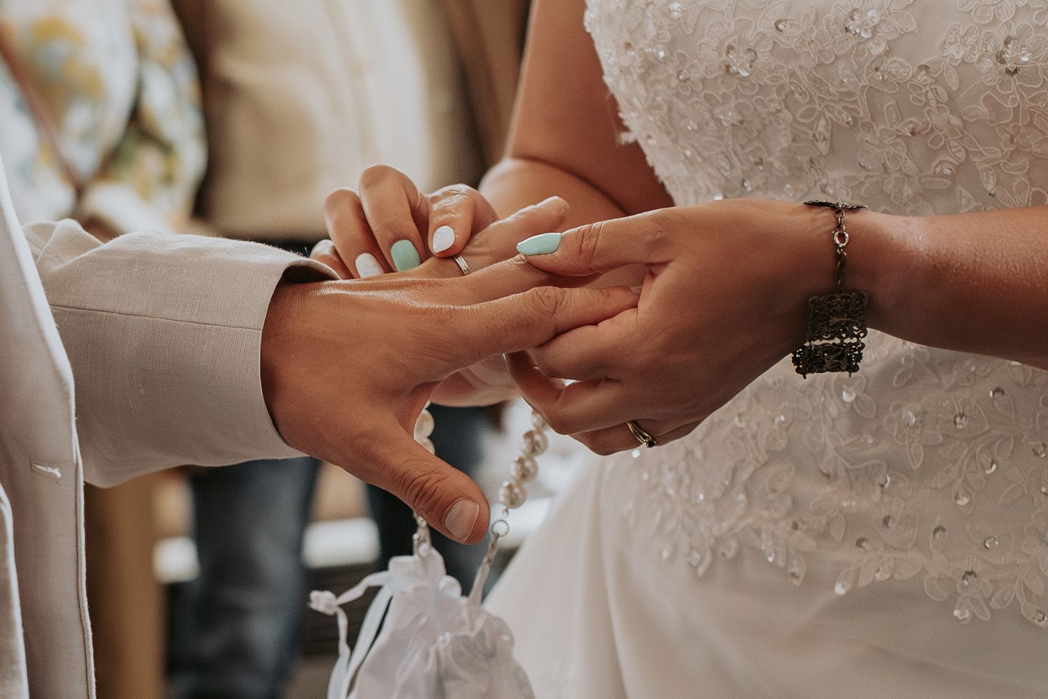Two hands exchanging wedding rings during an intimate ceremony with white dress and lace details in view.