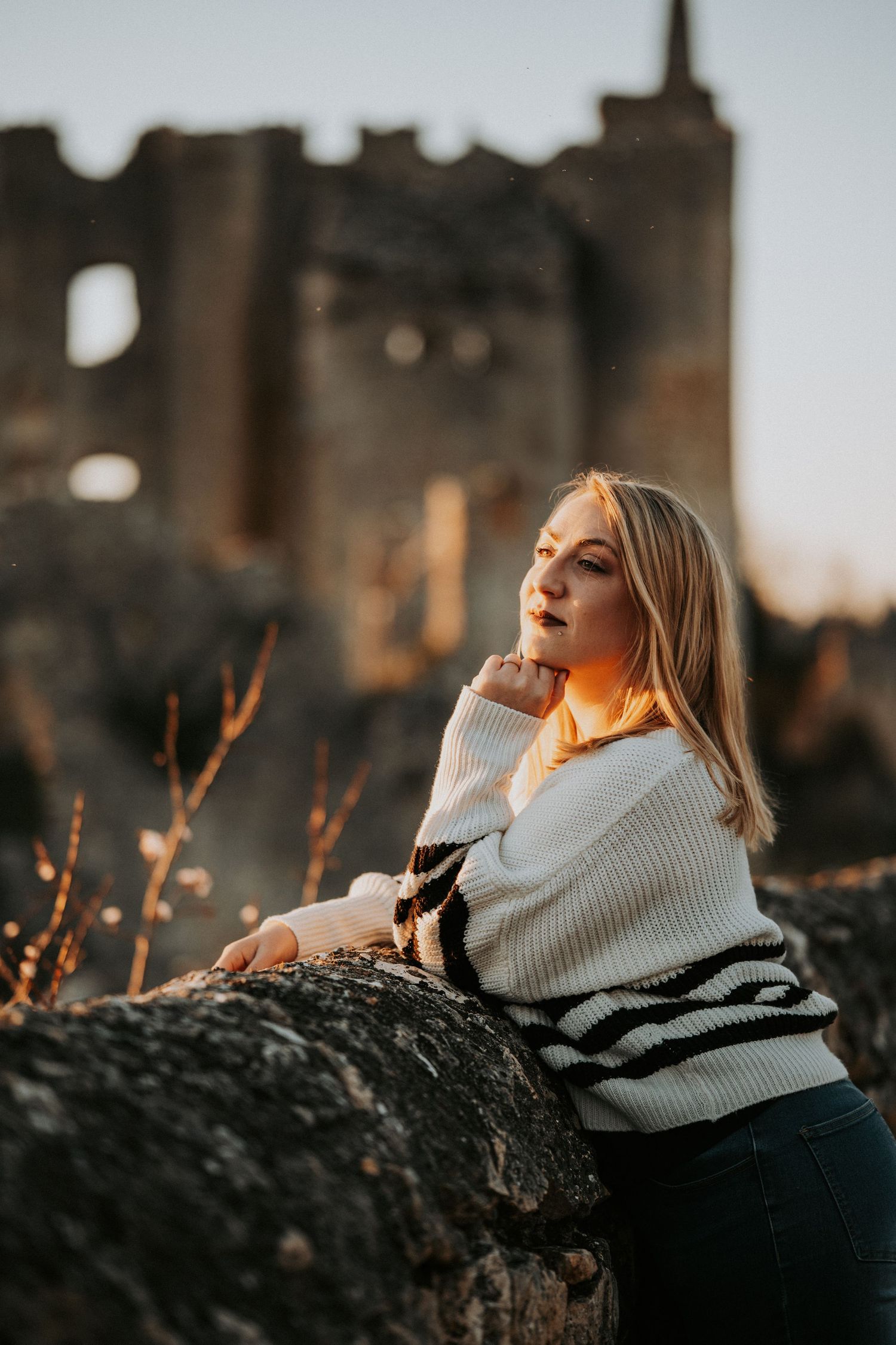 Blonde woman in white sweater poses against stone ruins at golden hour sunset.