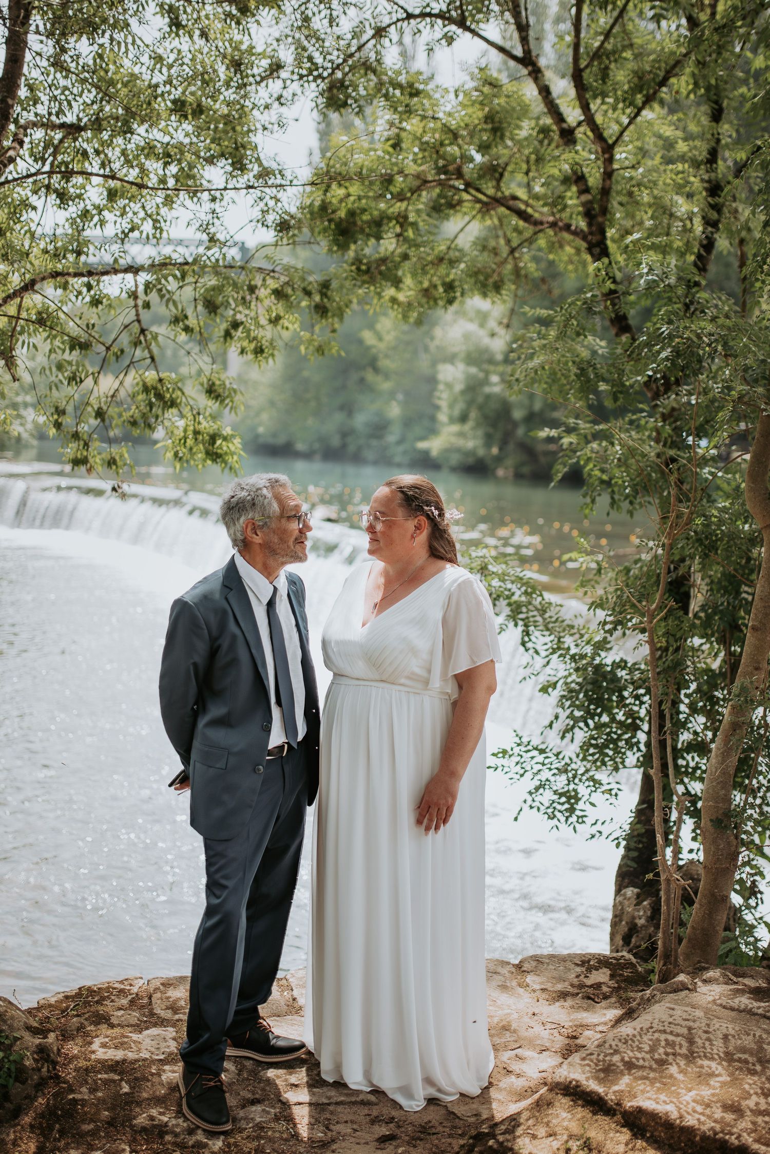 A couple shares a romantic moment by a serene lake with trees and a waterfall in the background.