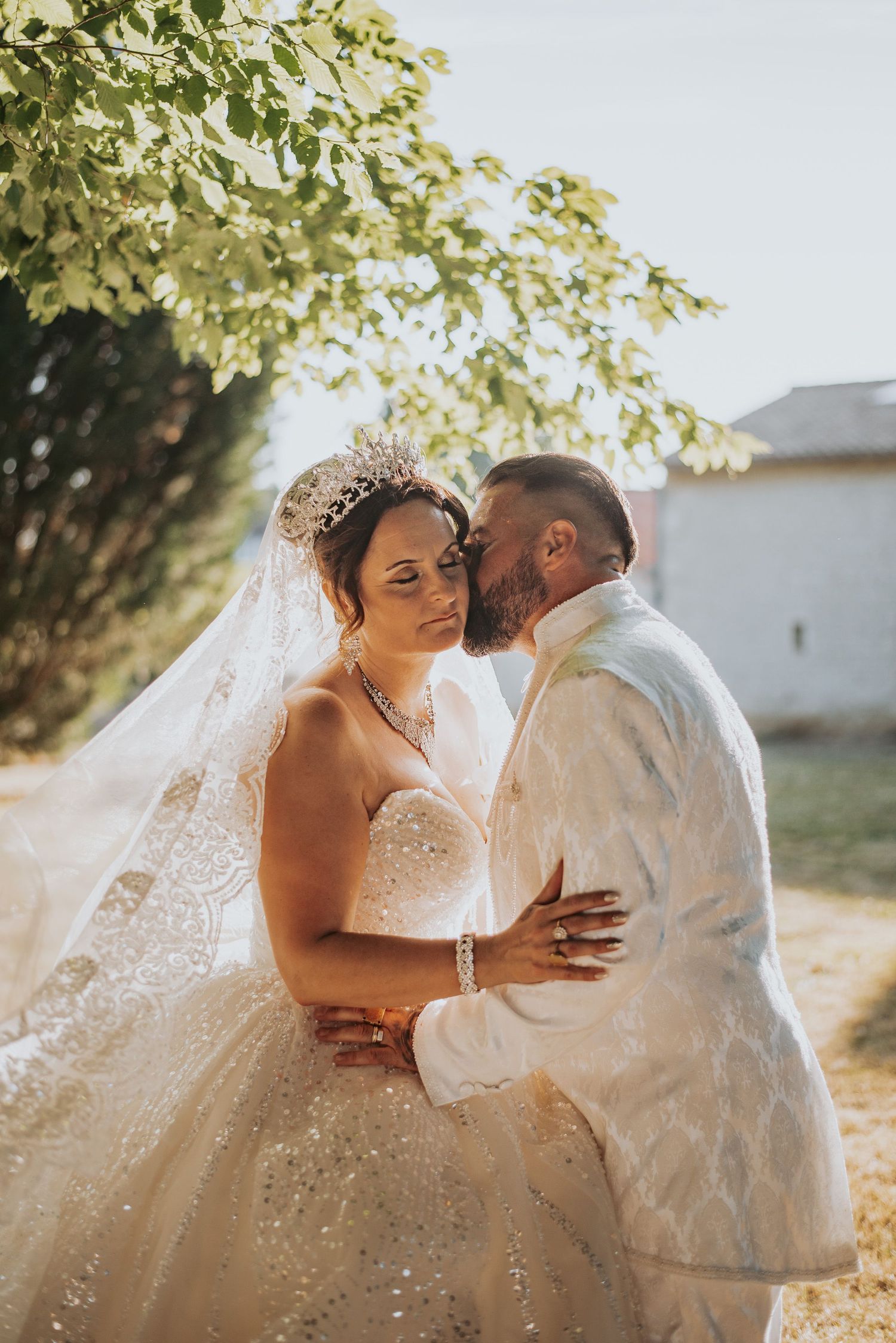 A romantic wedding couple shares tender moments in a sunlit outdoor setting with flowing white dress and veil.