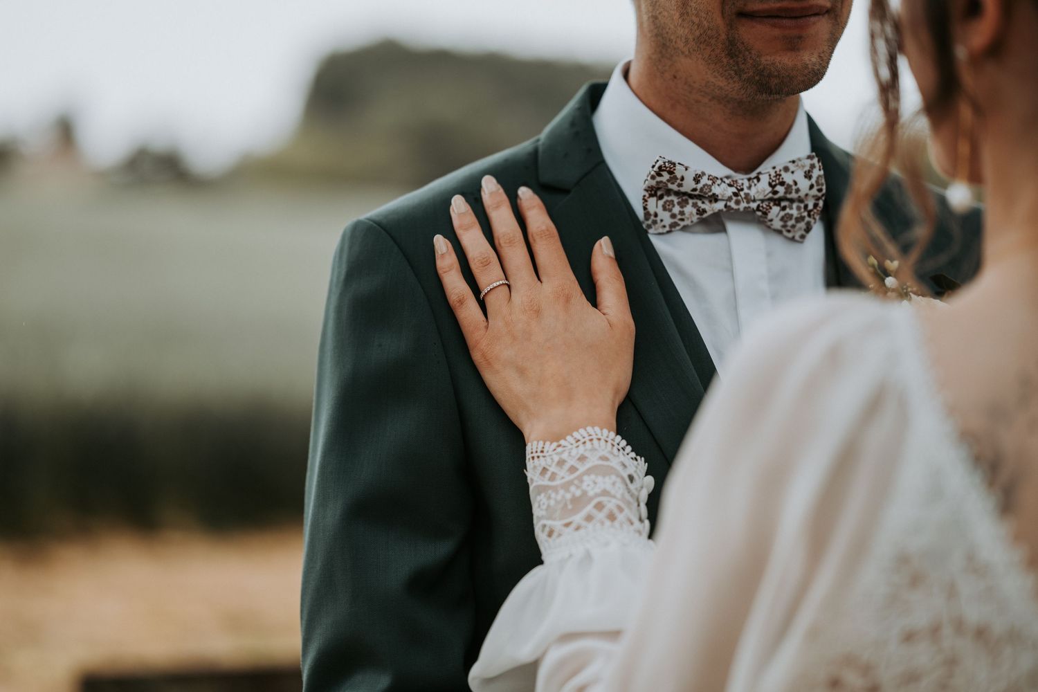 Bride in lace dress places hand on groom's chest, showcasing wedding ring, as he wears dark suit with floral bow tie.