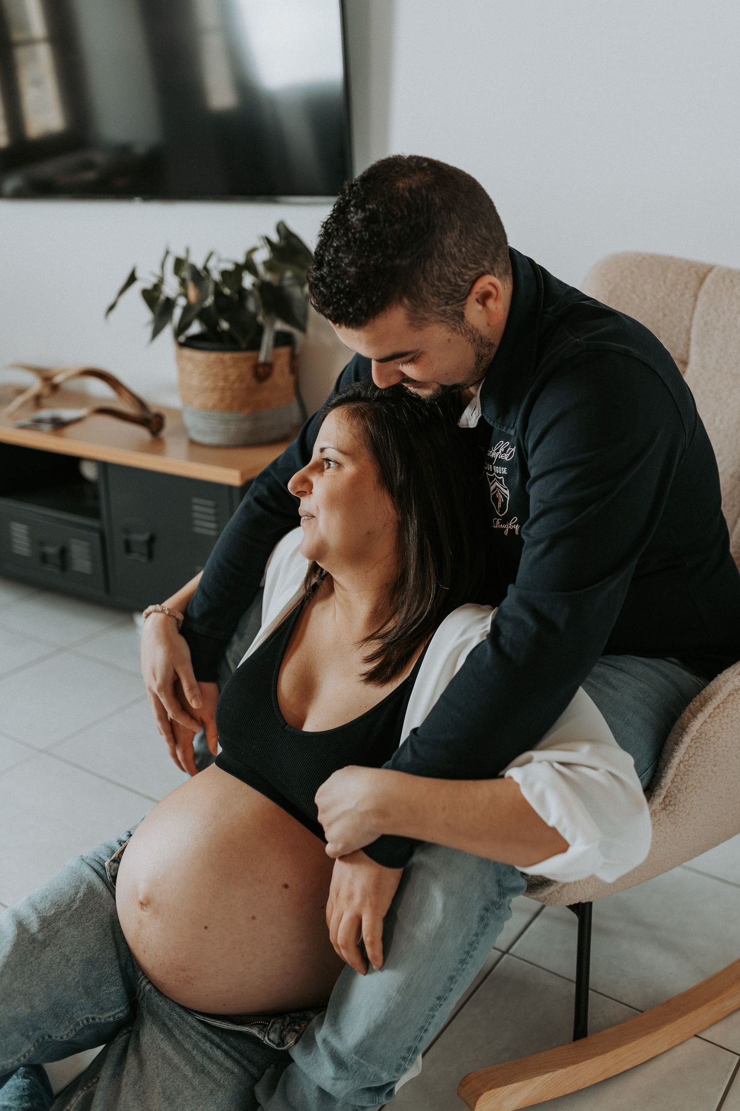 Intimate maternity photo of couple embracing in rocking chair at home with houseplant in background.