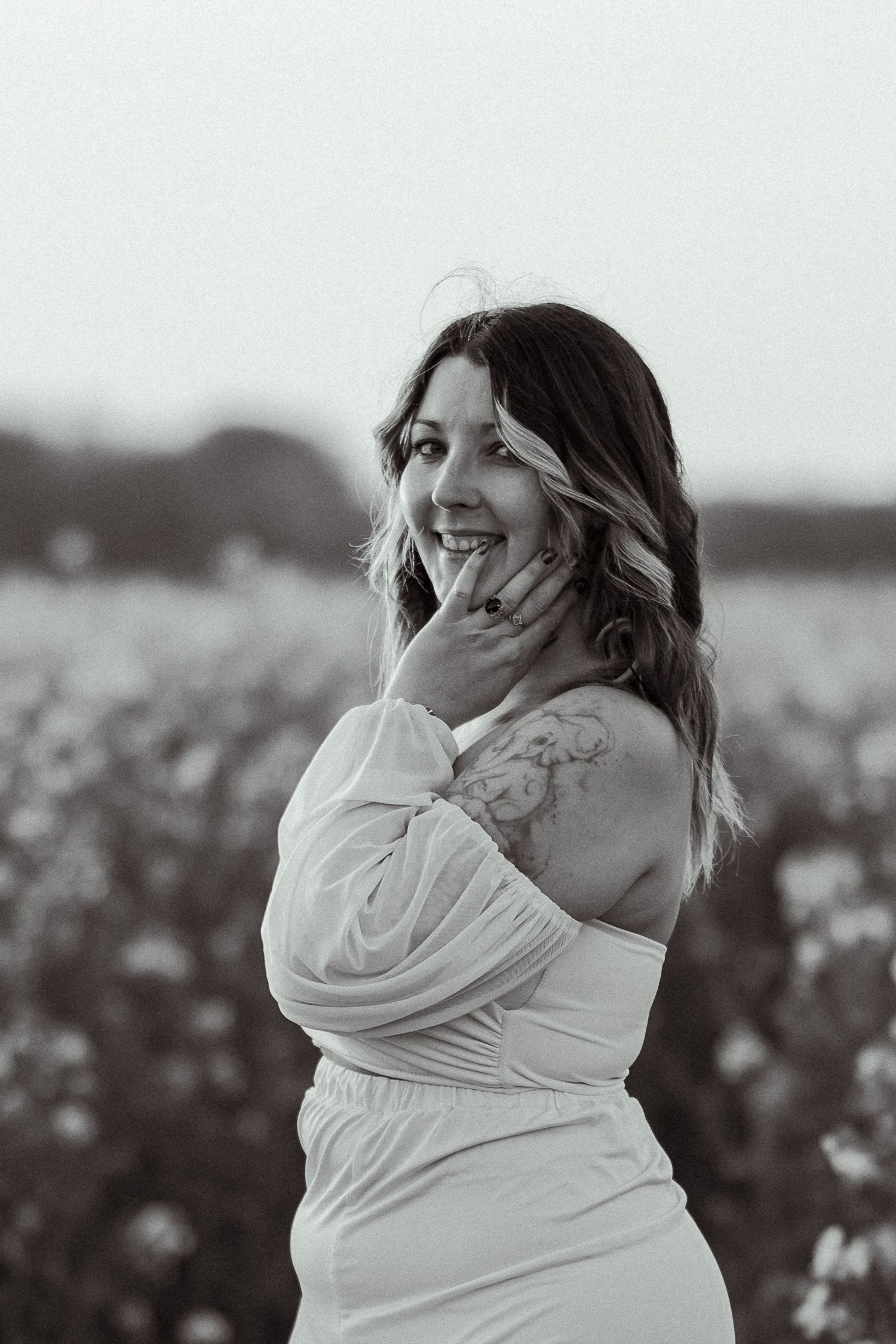 Black and white photo of a smiling woman in a white off-shoulder dress standing in a flower field.